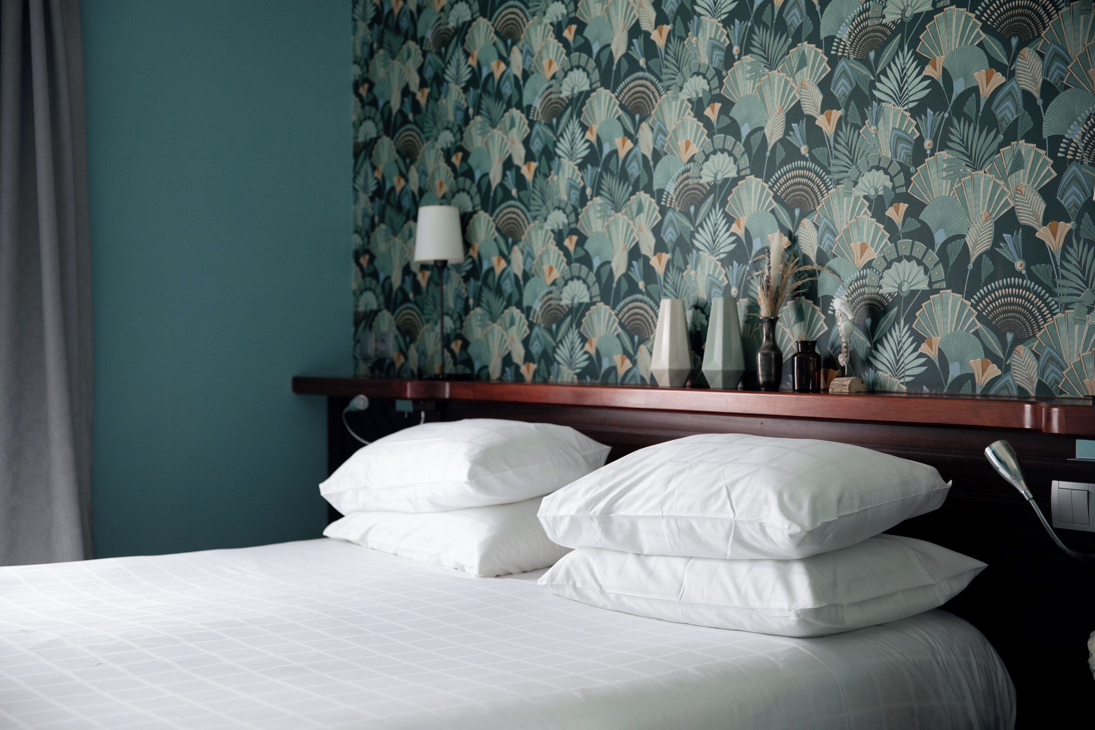 A neatly made bed with white linens and pillows, a wooden headboard, and a wall with a leafy pattern wallpaper.