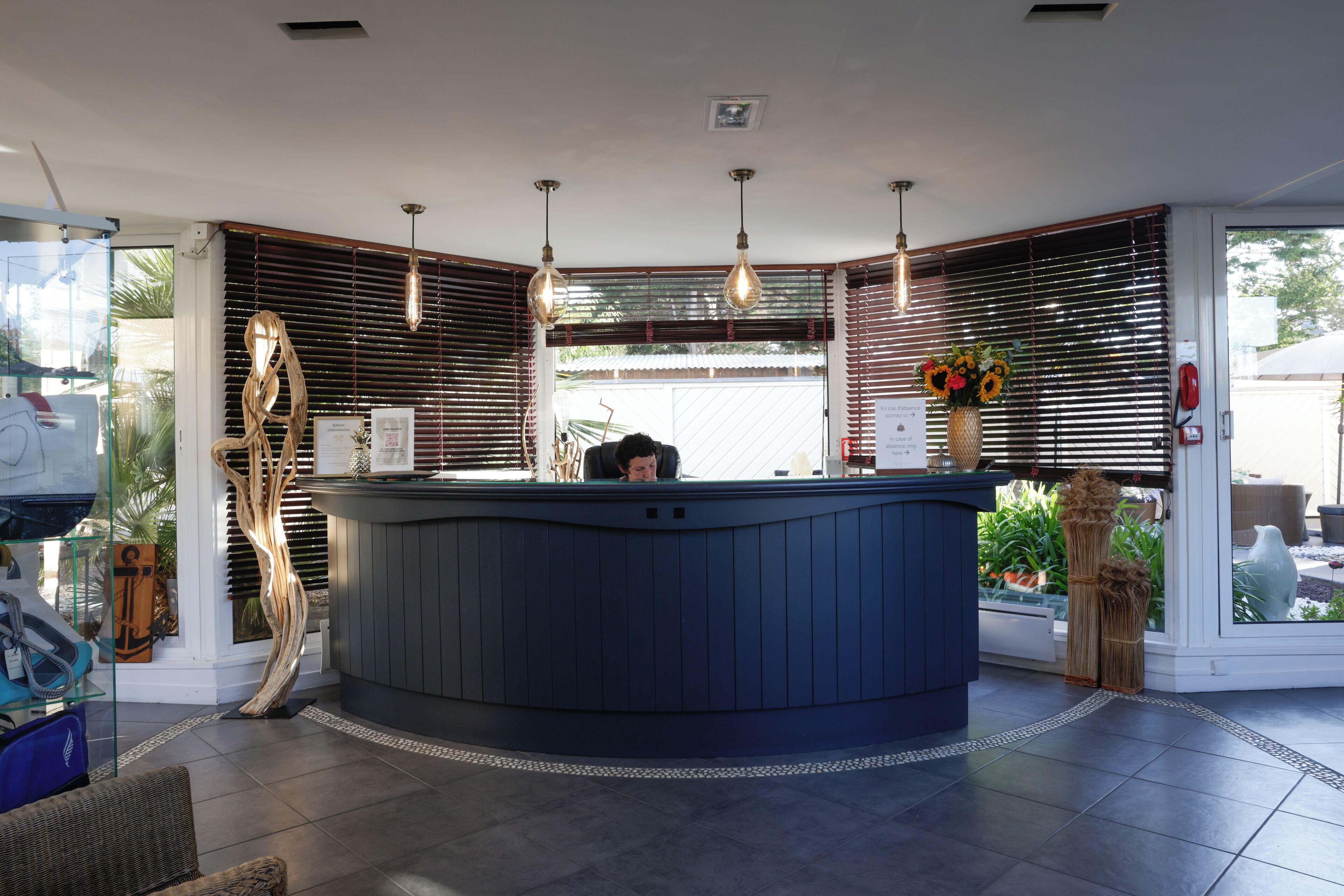 Una reception con una persona seduta al suo interno, in un ambiente moderno con persiane in legno, illuminazione a sospensione e vista sull'esterno.