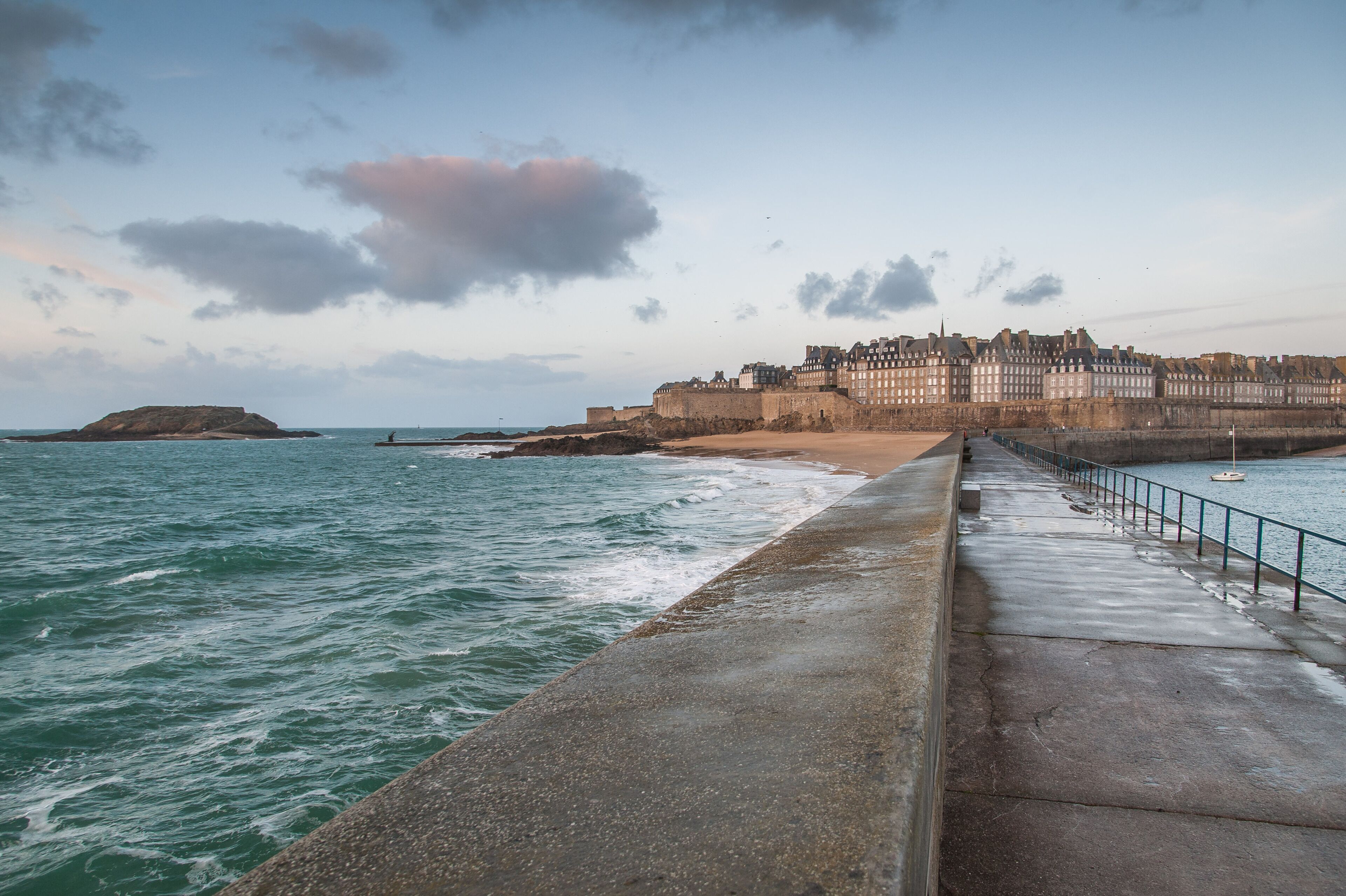 A coastal cityscape with a prominent castle-like structure, a promenade, and a calm sea.
