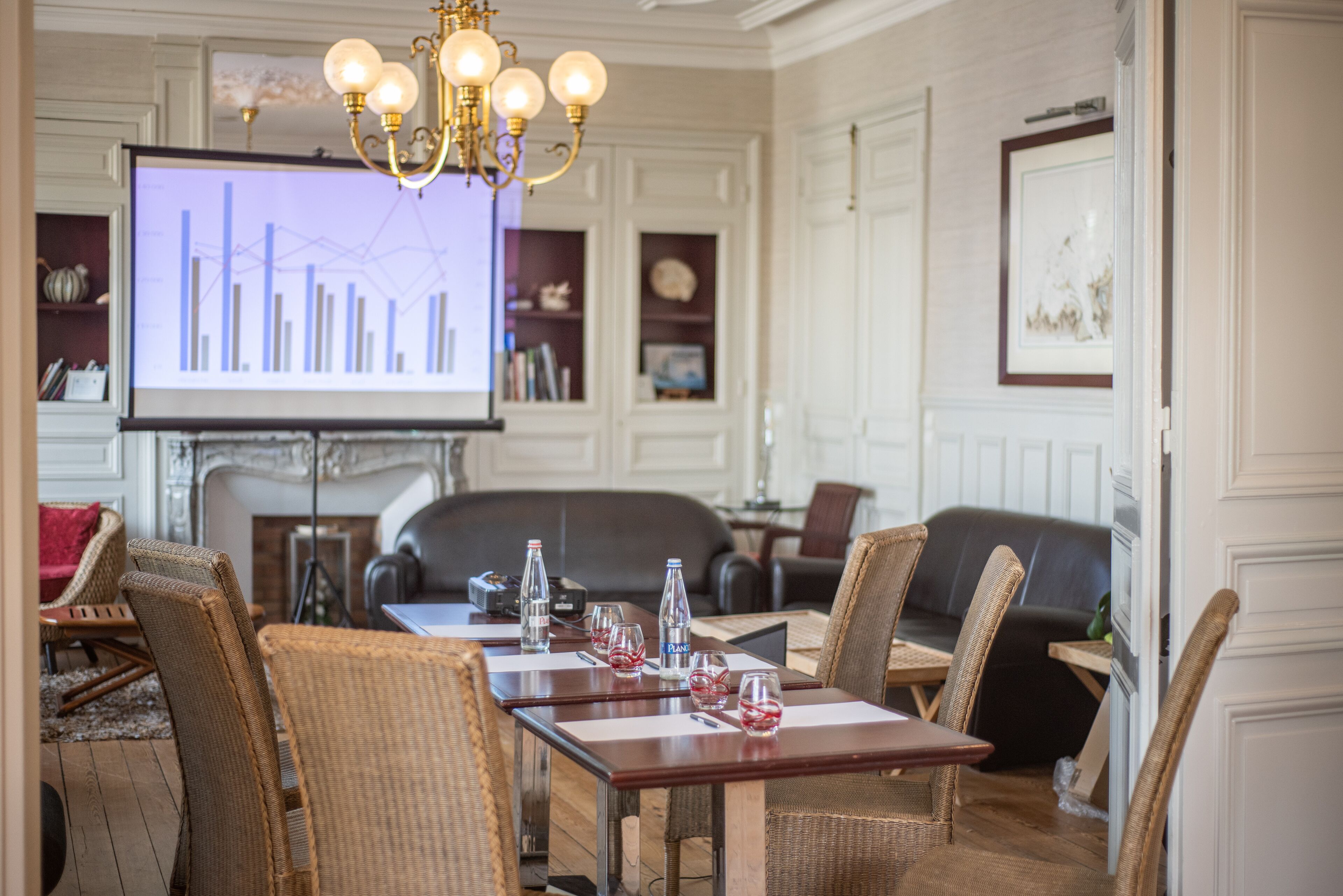 A conference room with a large screen displaying a graph, wicker chairs, a wooden table with water bottles, and a chandelier.