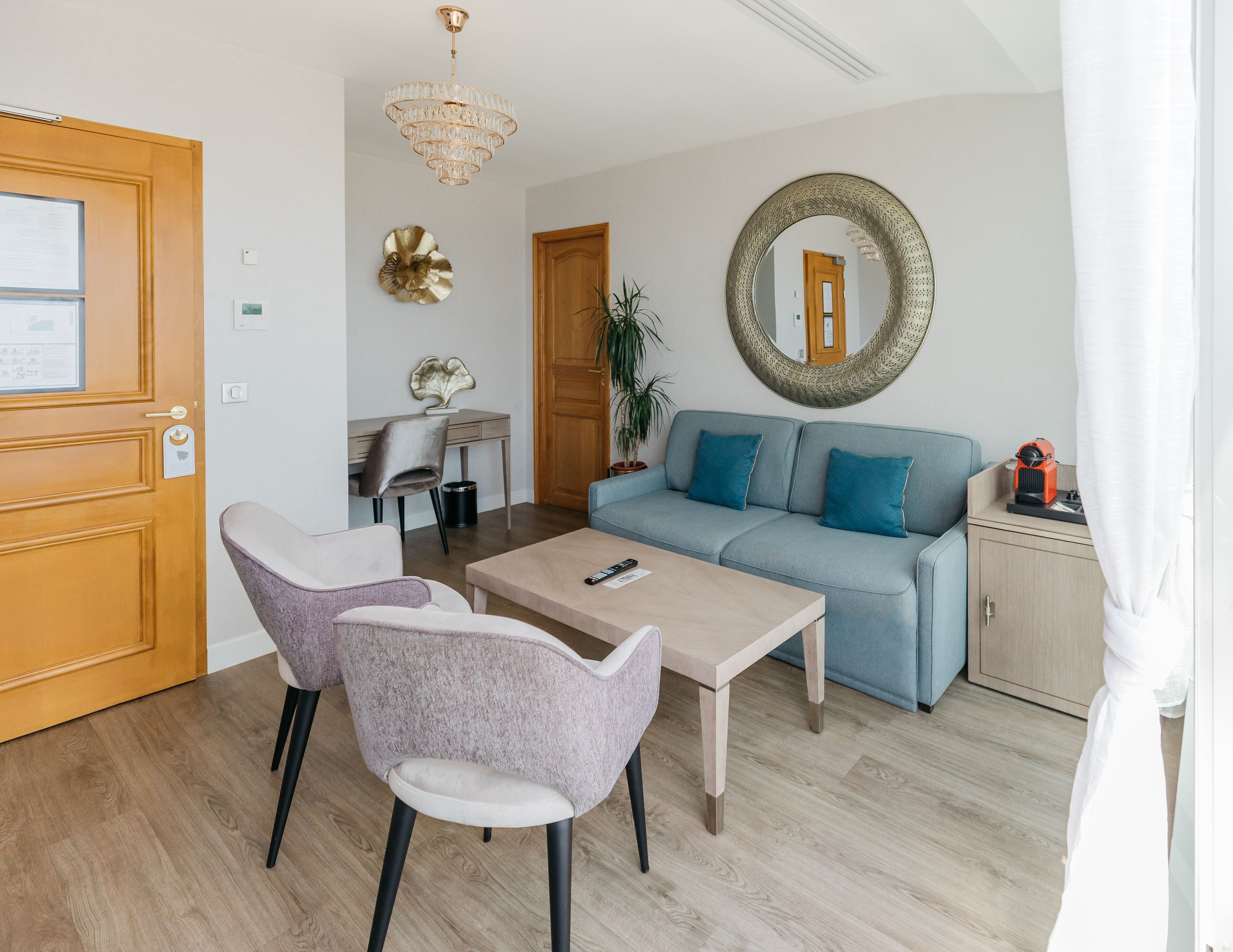 A modern living room with a sofa, two chairs, a coffee table, a chandelier, a round mirror, and a small cabinet.