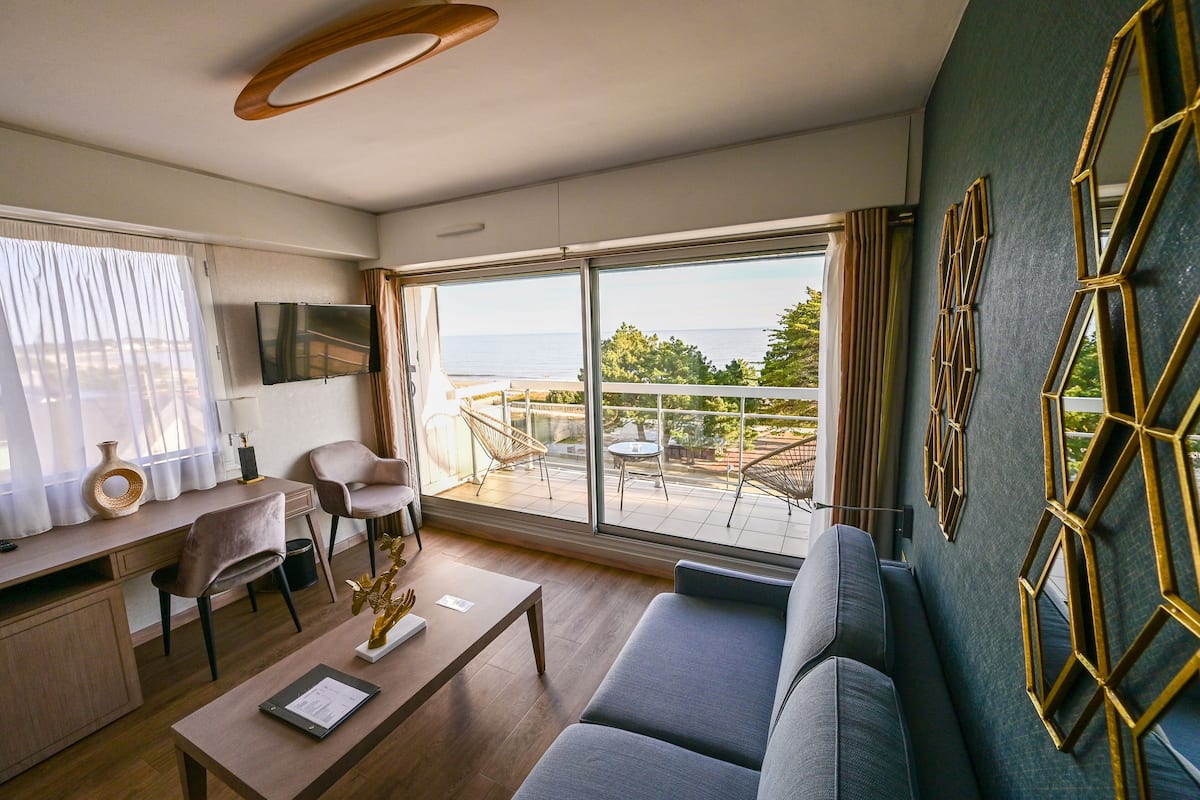 Ein modernes Wohnzimmer mit Sofa, Couchtisch, Schreibtisch und Fernseher. Ein Balkon bietet Blick auf das Meer und Bäume.