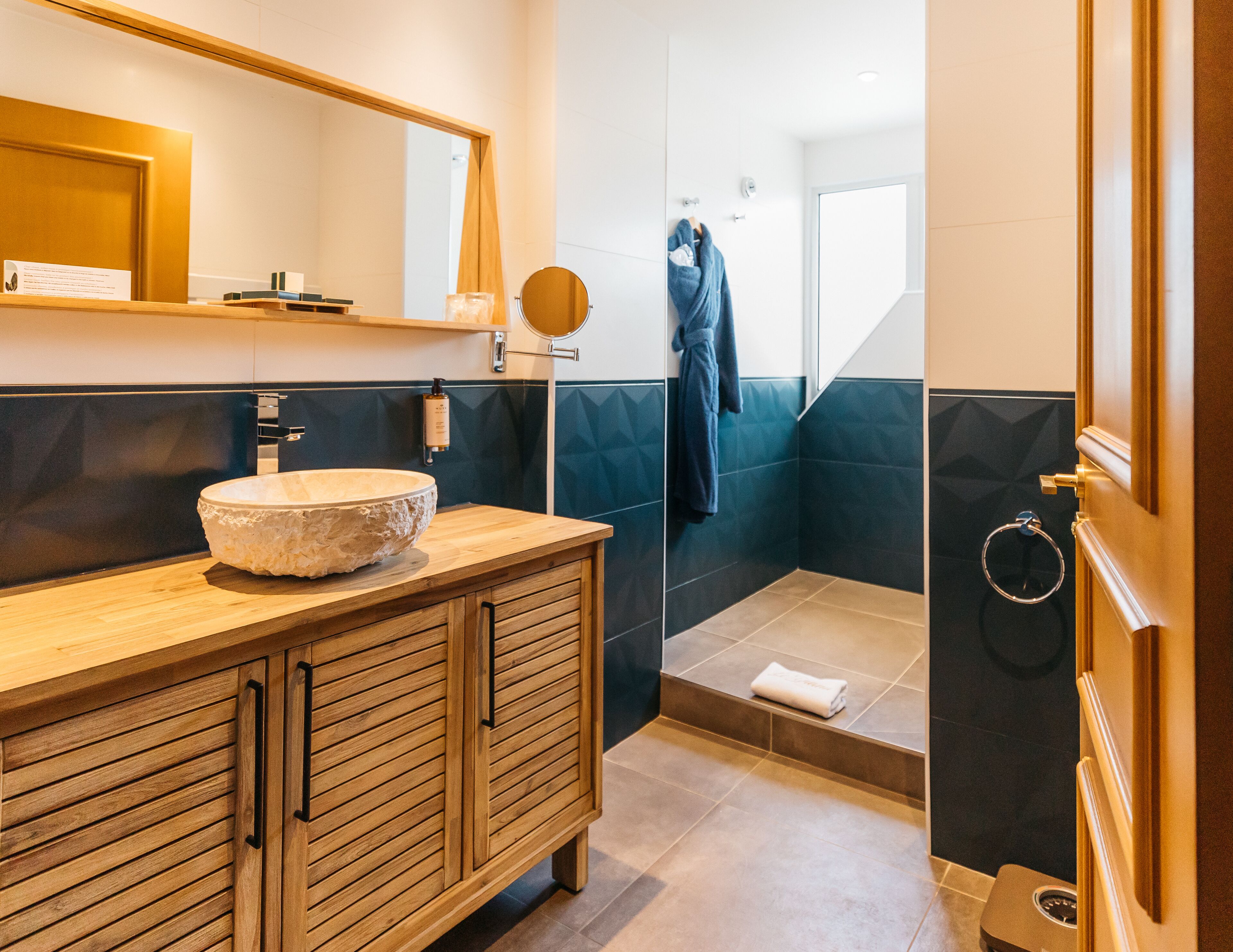 Un baño moderno con tocador de madera, un espejo redondo grande, un lavabo de piedra y una zona de ducha con aro para toallas.