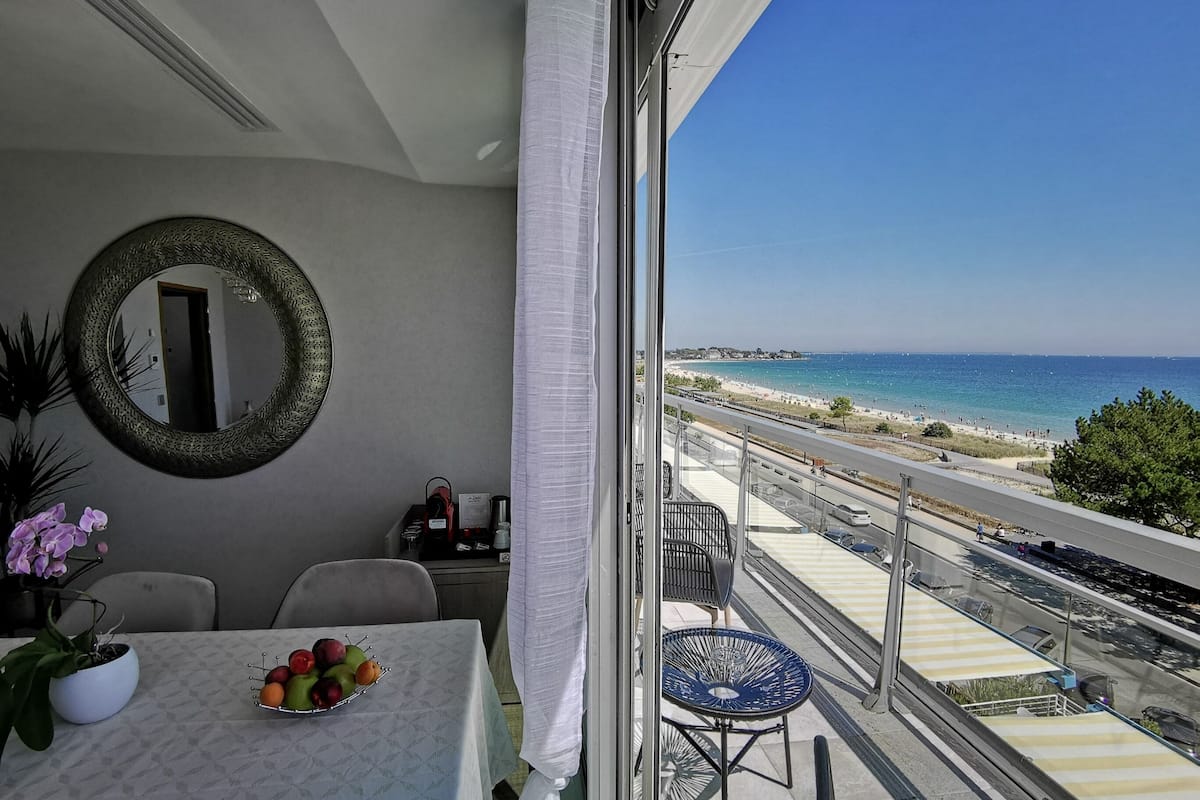 Ein Balkon mit Blick auf den Strand, ein Tisch mit Obst, ein Stuhl und ein Topfpflanze.