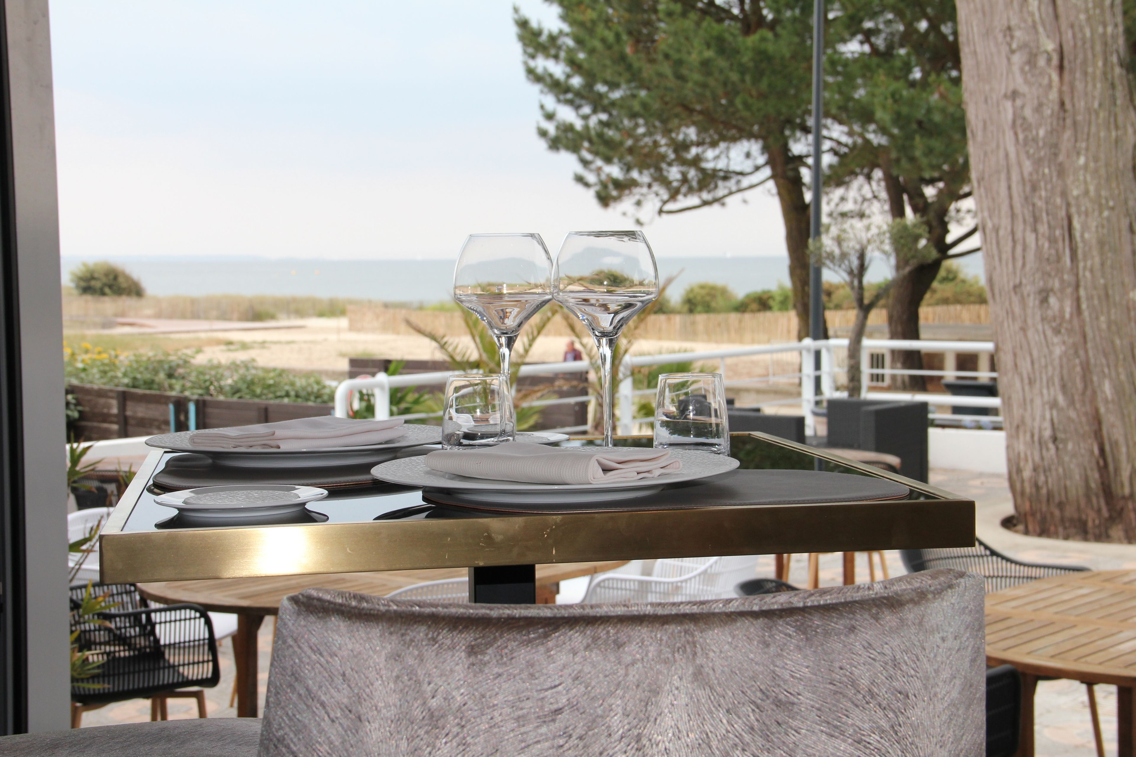 An outdoor dining area with a view of the beach, a table set with plates, glasses, and cutlery.