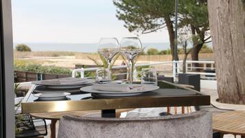 An outdoor dining area with a view of the beach, a table set with plates, glasses, and cutlery.