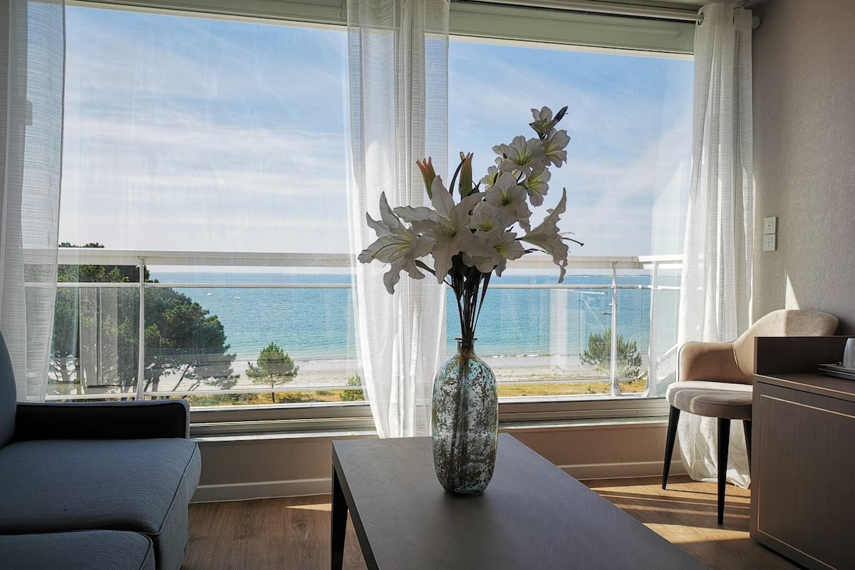 Ein modernes Wohnzimmer mit einem großen Fenster mit Blick auf einen Strand, einer Tischvase mit weißen Blumen sowie einem Sofa und einem Sessel.
