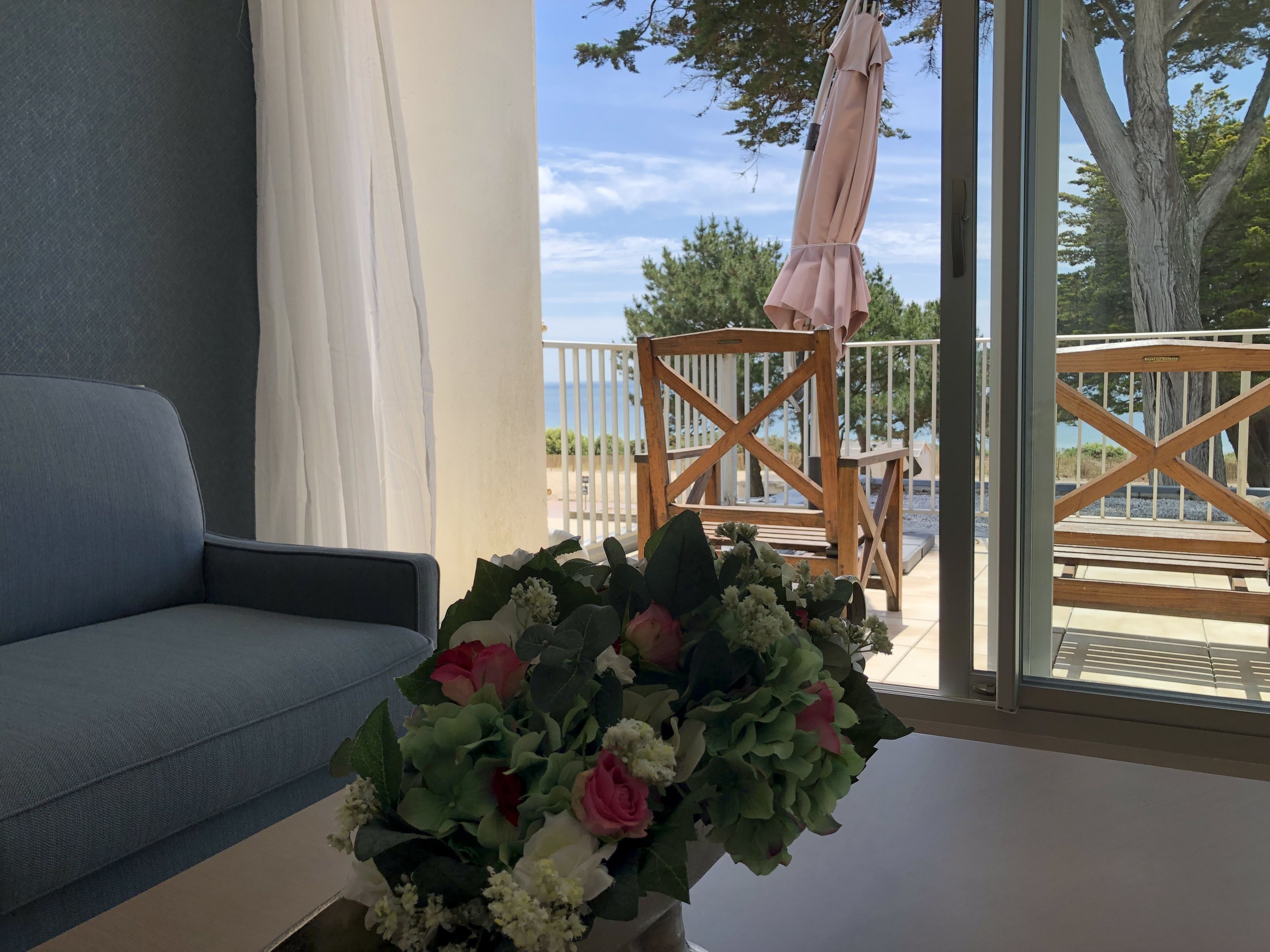 Un balcon avec une chaise en bois, une table agrémentée d’un arrangement floral et une vue sur la mer.