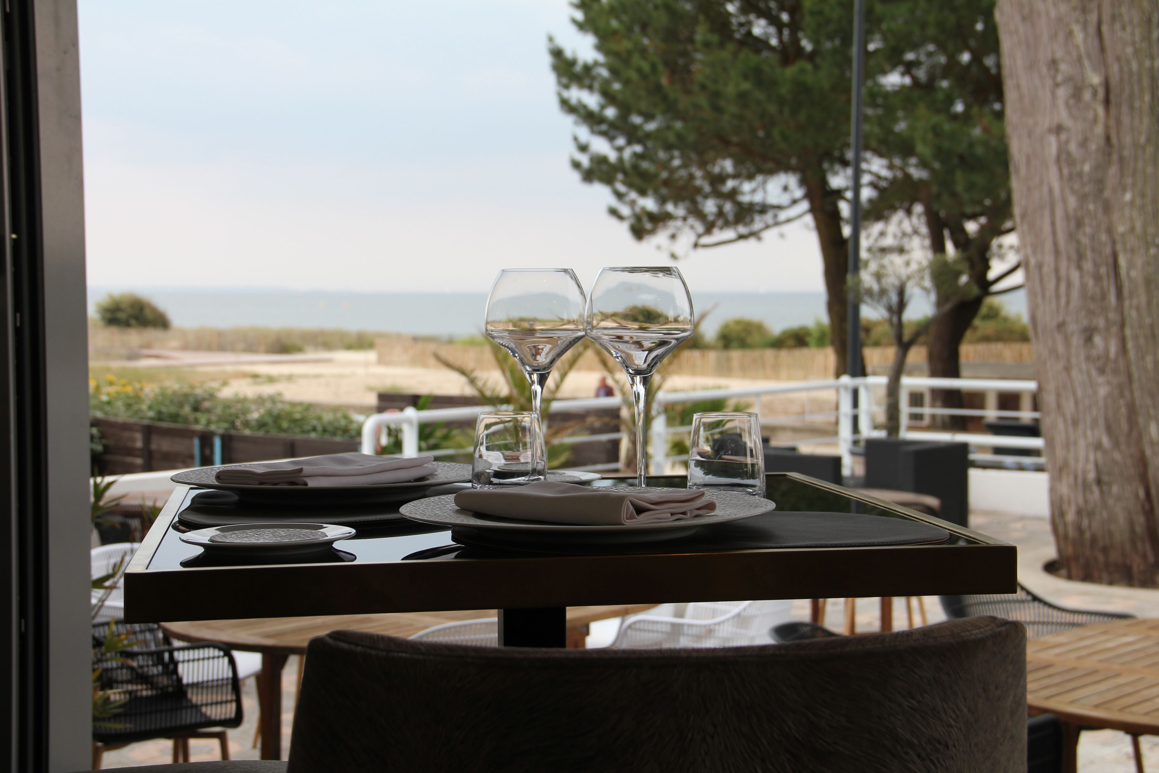 Een tafel gedekt voor twee personen, met wijnglazen, borden en bestek, met uitzicht op een strand.