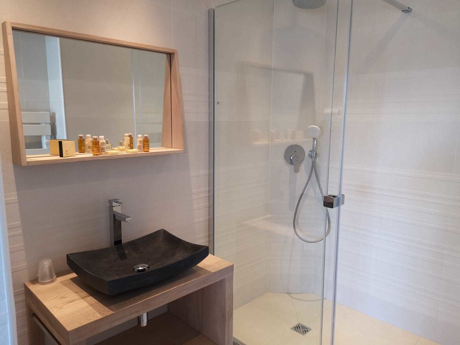 Een moderne badkamer met een glazen douchecabine, een houten wastafelmeubel met een zwarte wastafel en een spiegel erboven.