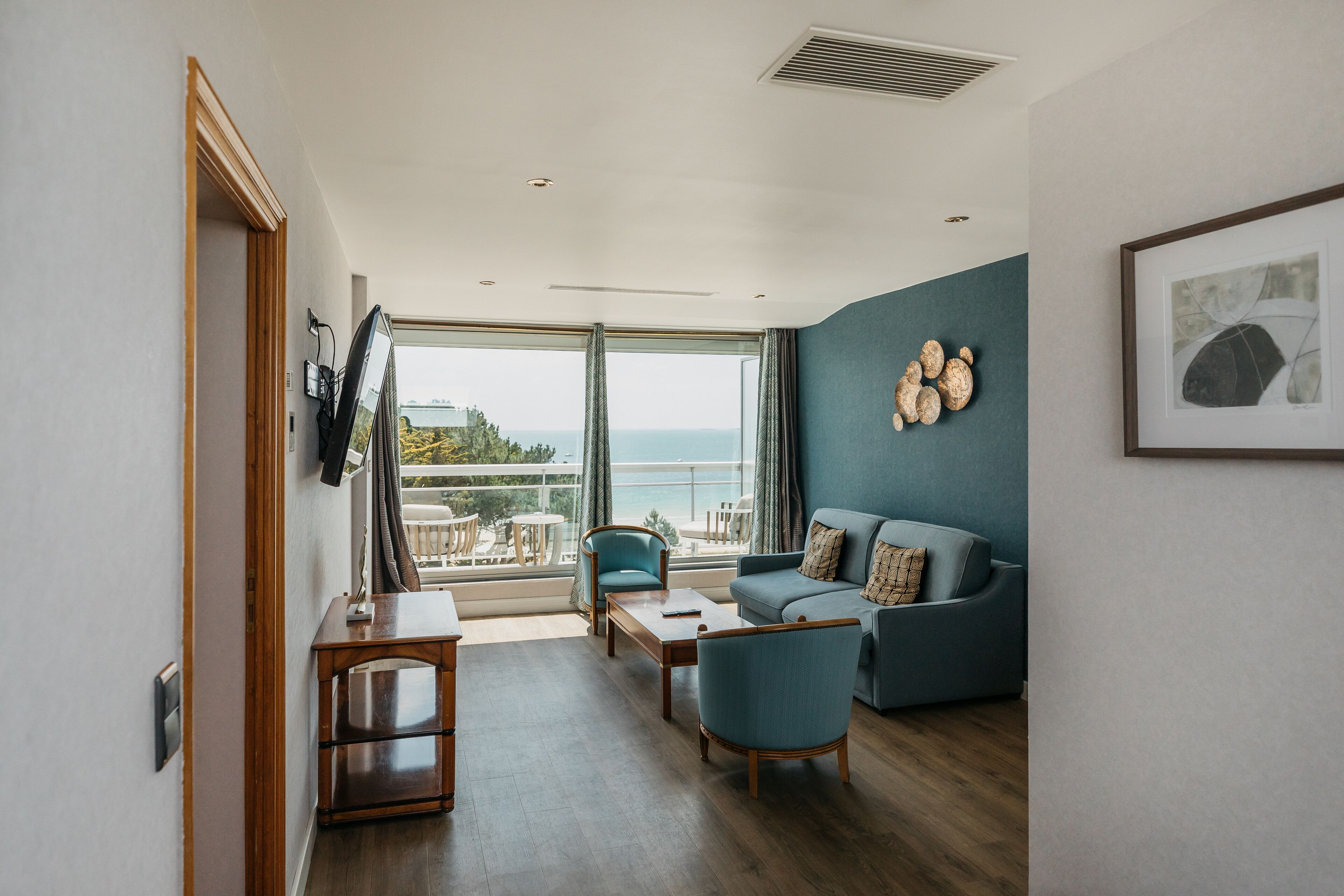 A modern living room with a sofa, chairs, and a table. There is a large window with a view of the sea.
