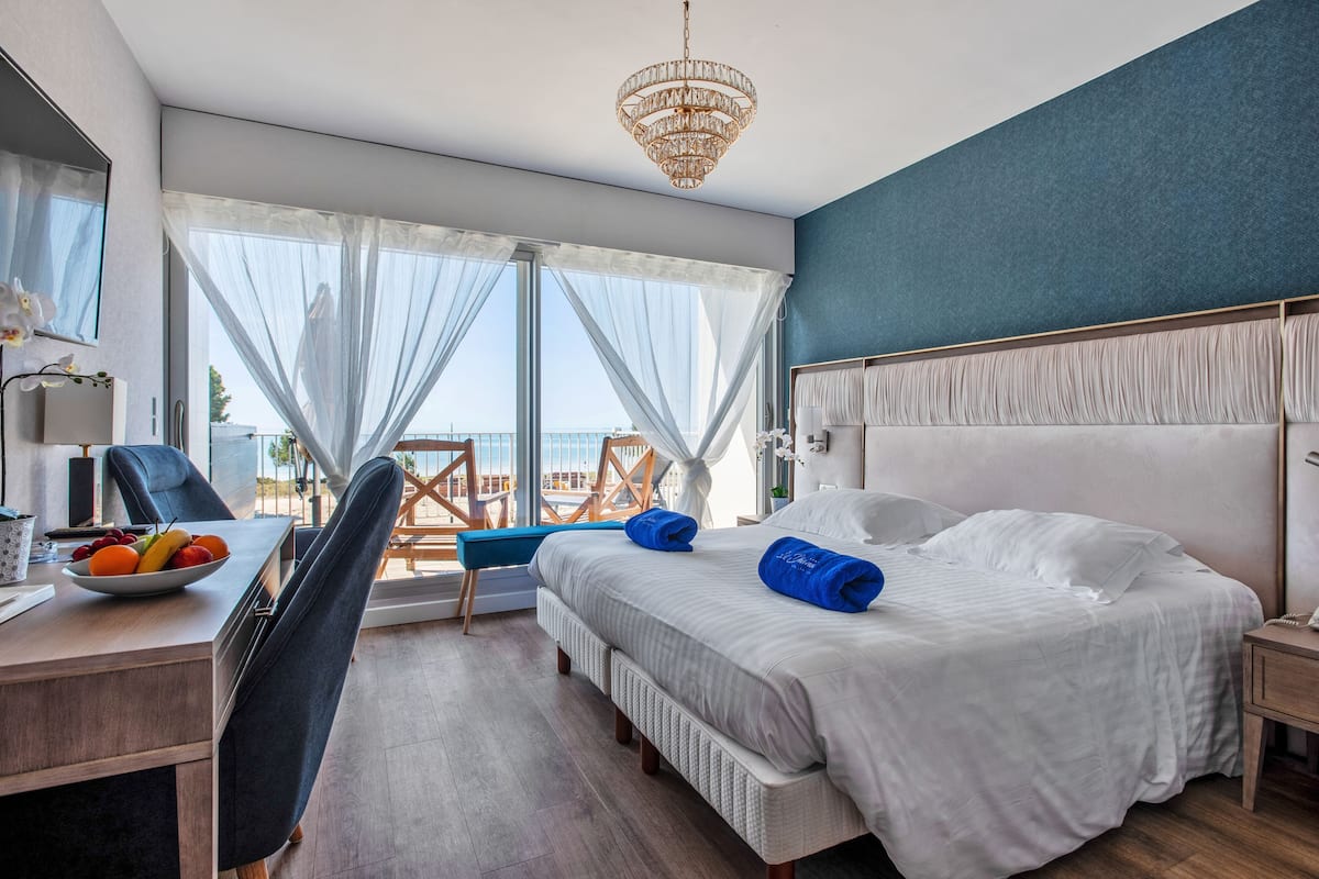 Ein modernes Hotelzimmer mit einem großen Bett, einem Schreibtisch mit Obstschale, einem Sessel und Meerblick durch leichte Vorhänge.