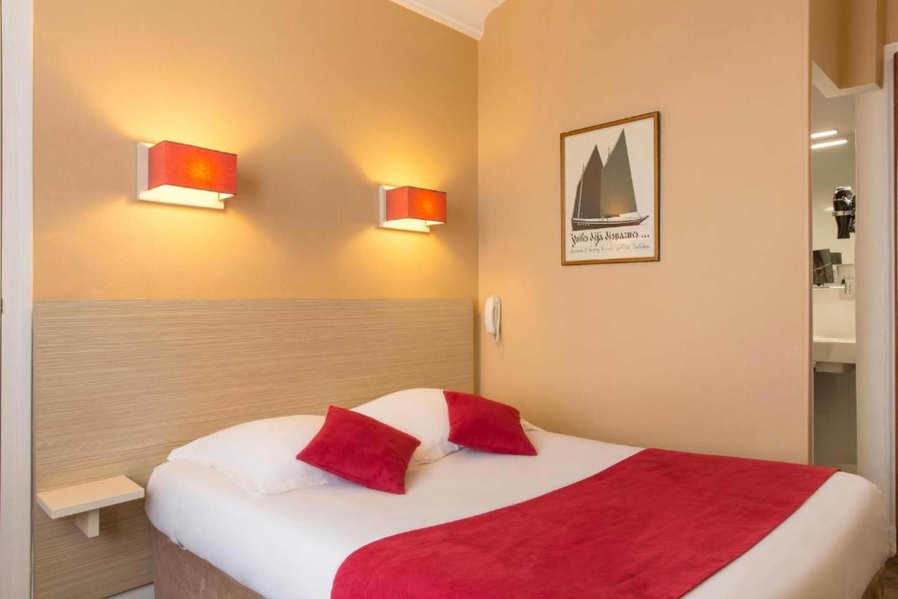 A hotel room with a bed, two bedside lamps, a red throw blanket, and a sailboat picture on the wall.