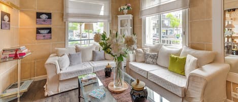 Un salón luminoso con paredes color beige, un conjunto de sofá blanco, una mesa de centro de vidrio y un jarrón con flores.
