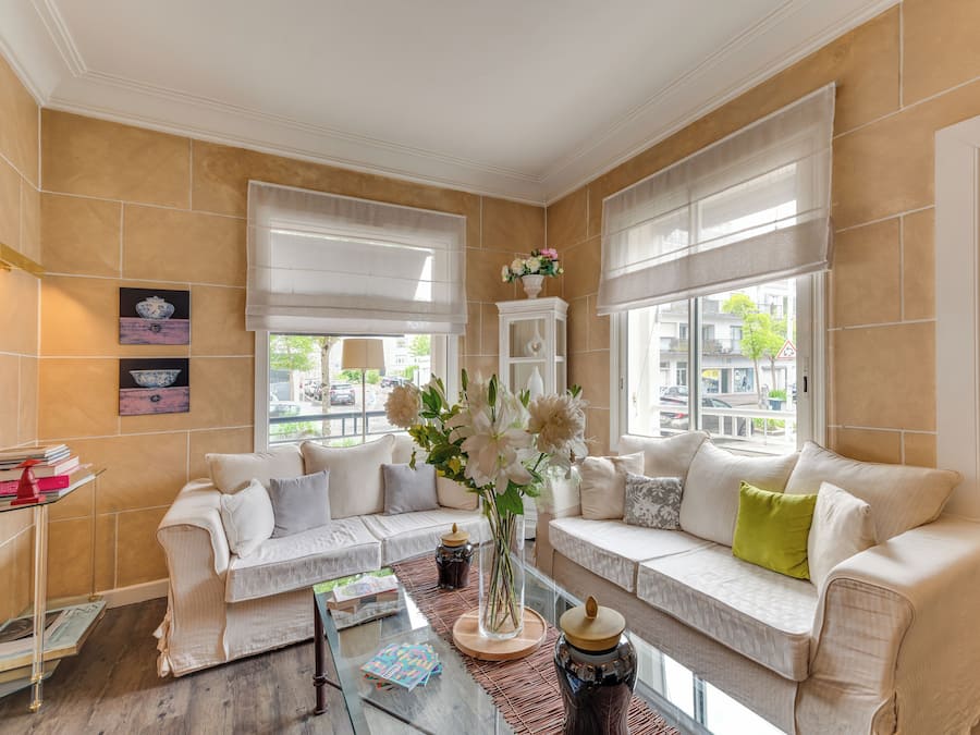 A well-lit living room with beige walls, a white sofa set, a glass coffee table, and a vase with flowers.