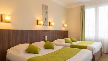 A hotel room with two beds, each with a green pillow and blanket, a wooden headboard, and wall-mounted lights.