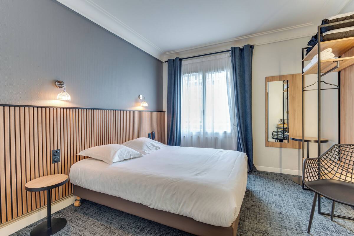 A hotel room with a bed, a small table, a chair, a mirror, and a window with curtains.
