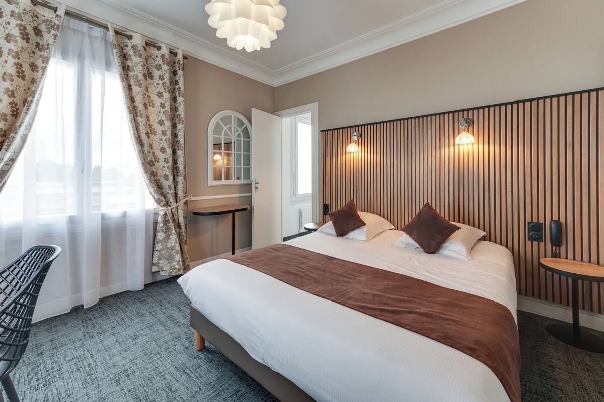 A hotel room with a large bed, a desk, a chair, and a window with curtains.