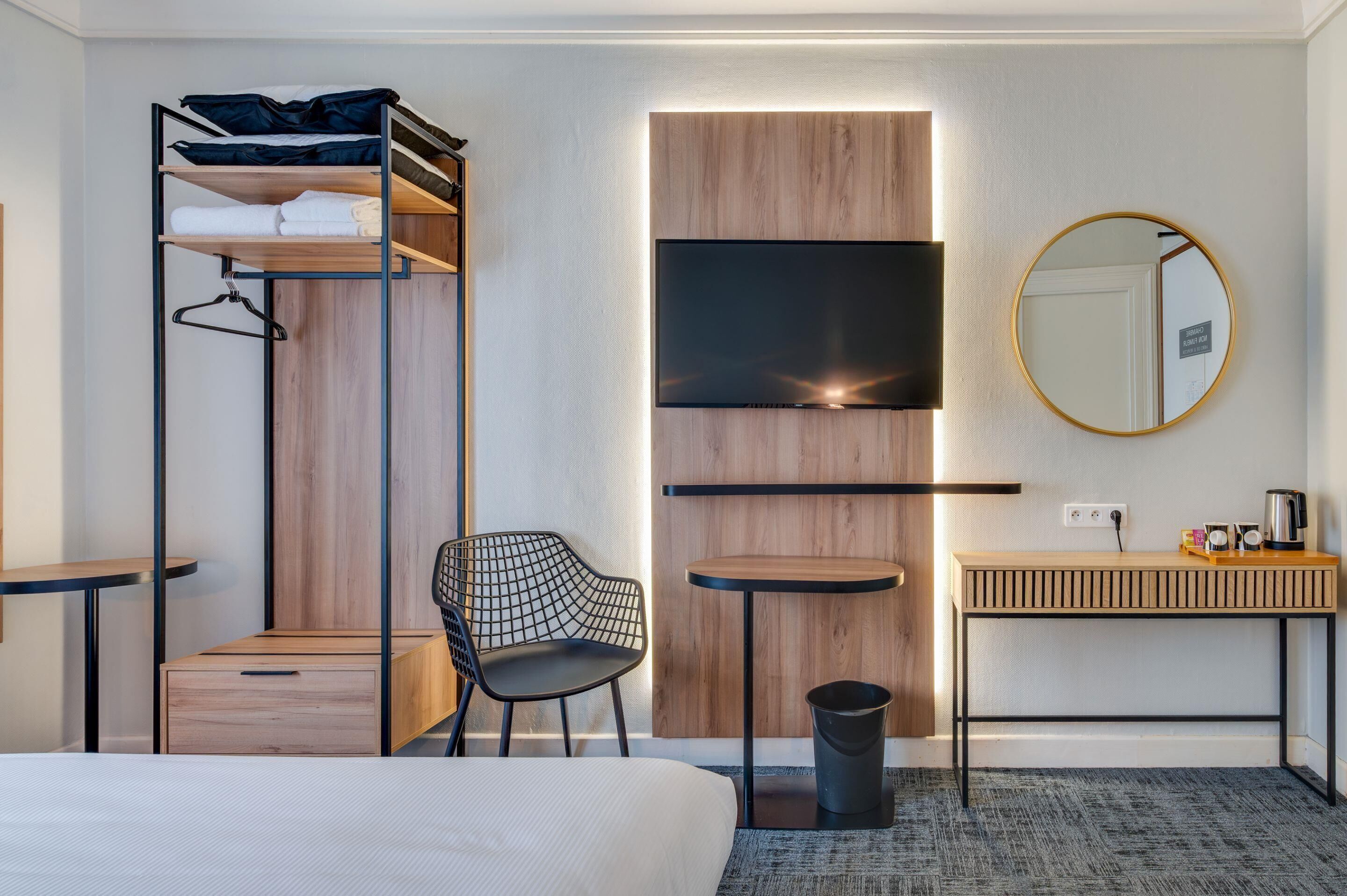 A hotel room with a bunk bed, a single bed, a chair, a small table, a mirror, a TV, and a bedside table with a kettle.
