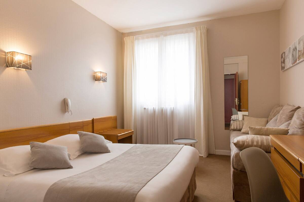 A hotel room with a large bed, a desk, a chair, a small table, and a window with curtains.