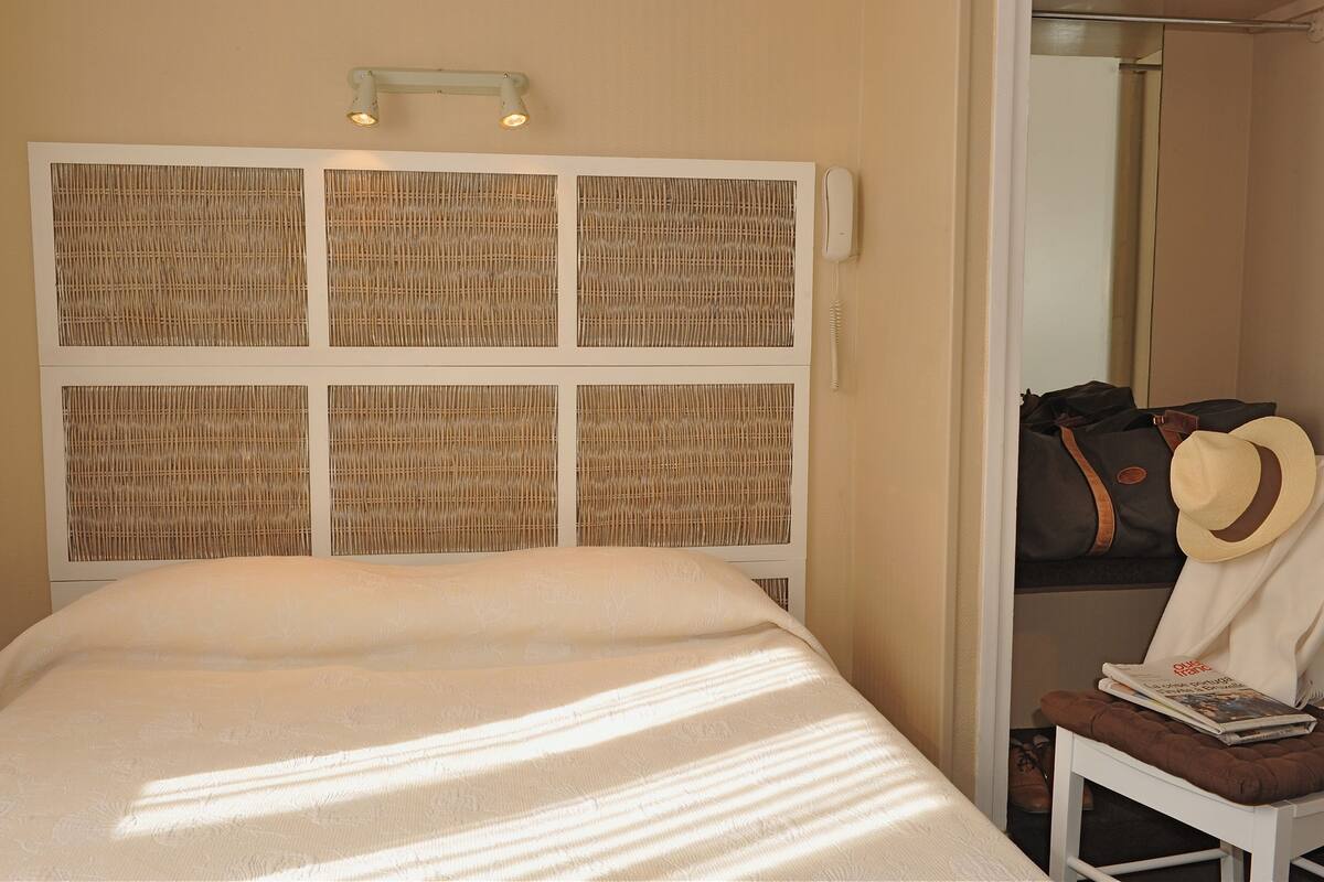 A neatly made bed with a white bedspread, a headboard with woven panels, a bedside lamp, and a small table with a hat and a magazine.