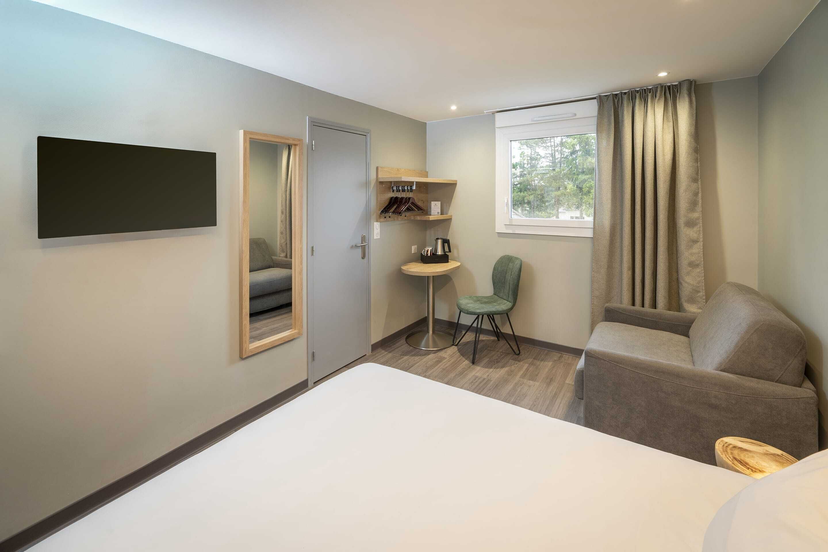 A hotel room with a bed, a seating area with a chair, a small table, a TV mounted on the wall, a window with curtains, and a coat rack with hangers.