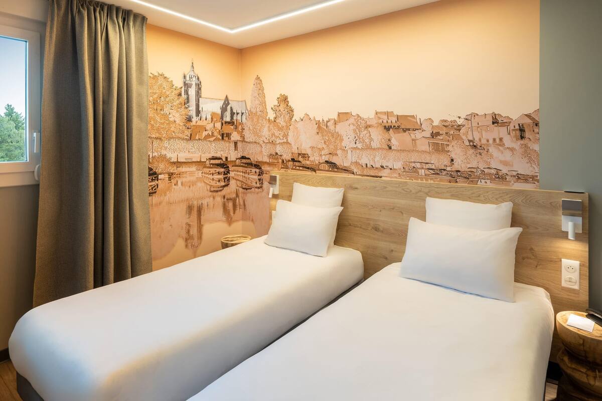 A hotel room with two beds, a cityscape mural on the wall, and a window with curtains.