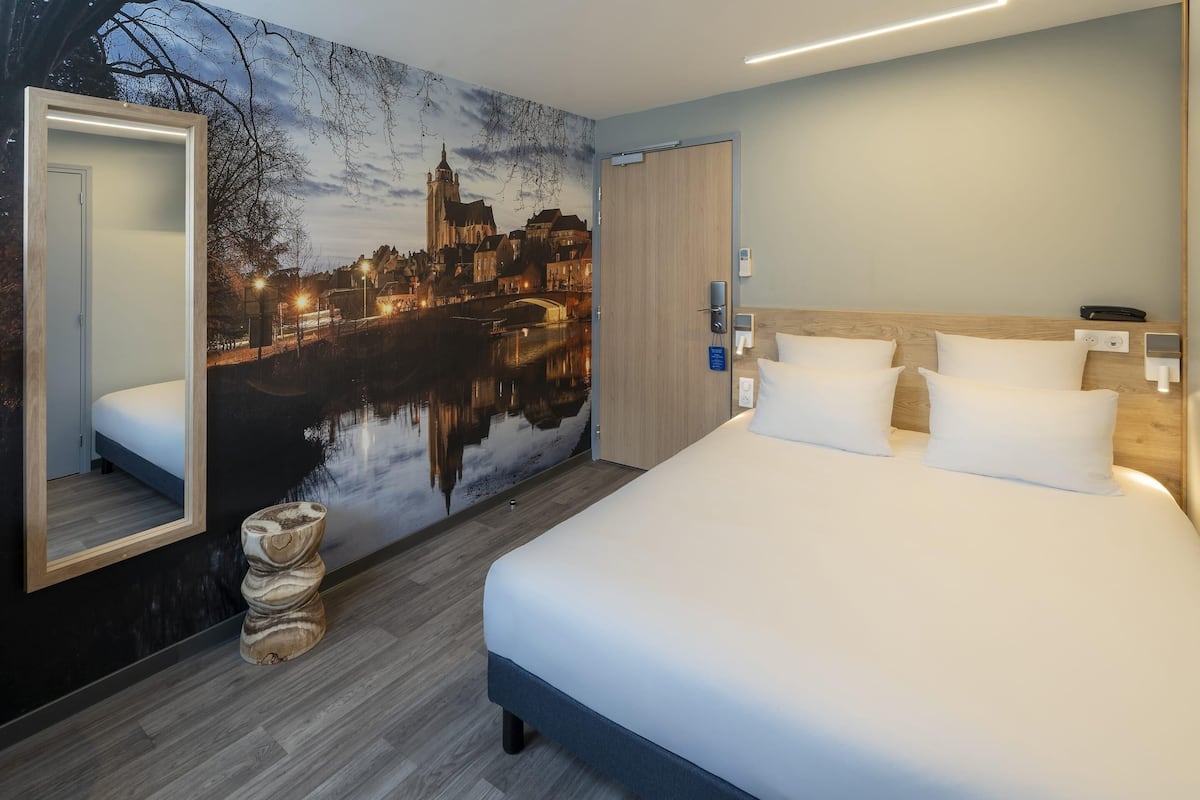 A modern hotel room with a large bed, a nightstand, a mirror, and a cityscape mural on the wall.