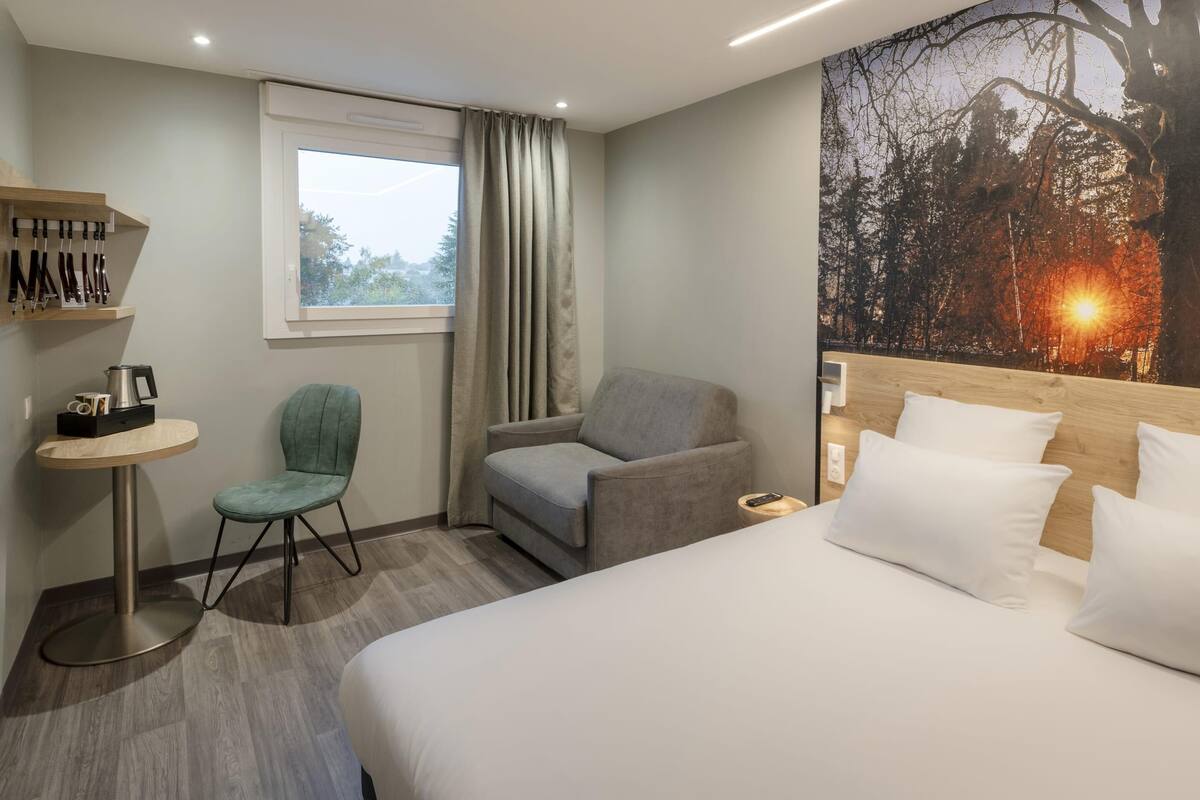 A hotel room with a bed, two armchairs, a small table with a kettle, a shelf with hanging items, a window with curtains, and a wall with a nature scene.