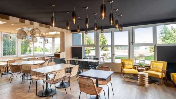 A spacious dining area with high tables and wicker chairs, large windows offering a view of the outdoors, and hanging pendant lights. - Sure Hotel by Best Western Dole (Dole)