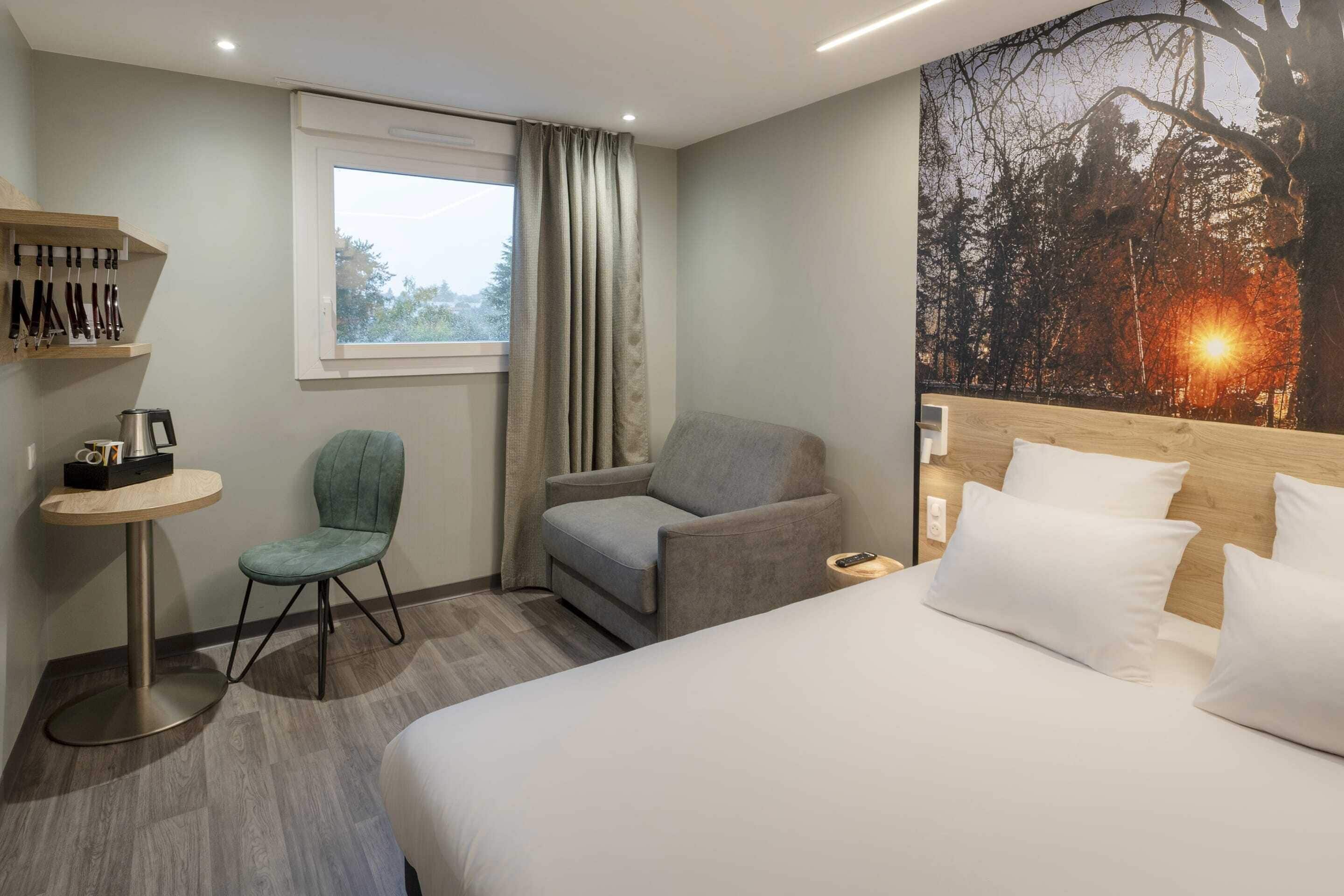 Habitación de hotel con cama, dos sillas, una mesita, ventana con cortinas y un cuadro con una escena de naturaleza.