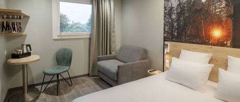 Habitación de hotel con cama, dos sillas, una mesita, ventana con cortinas y un cuadro con una escena de naturaleza.