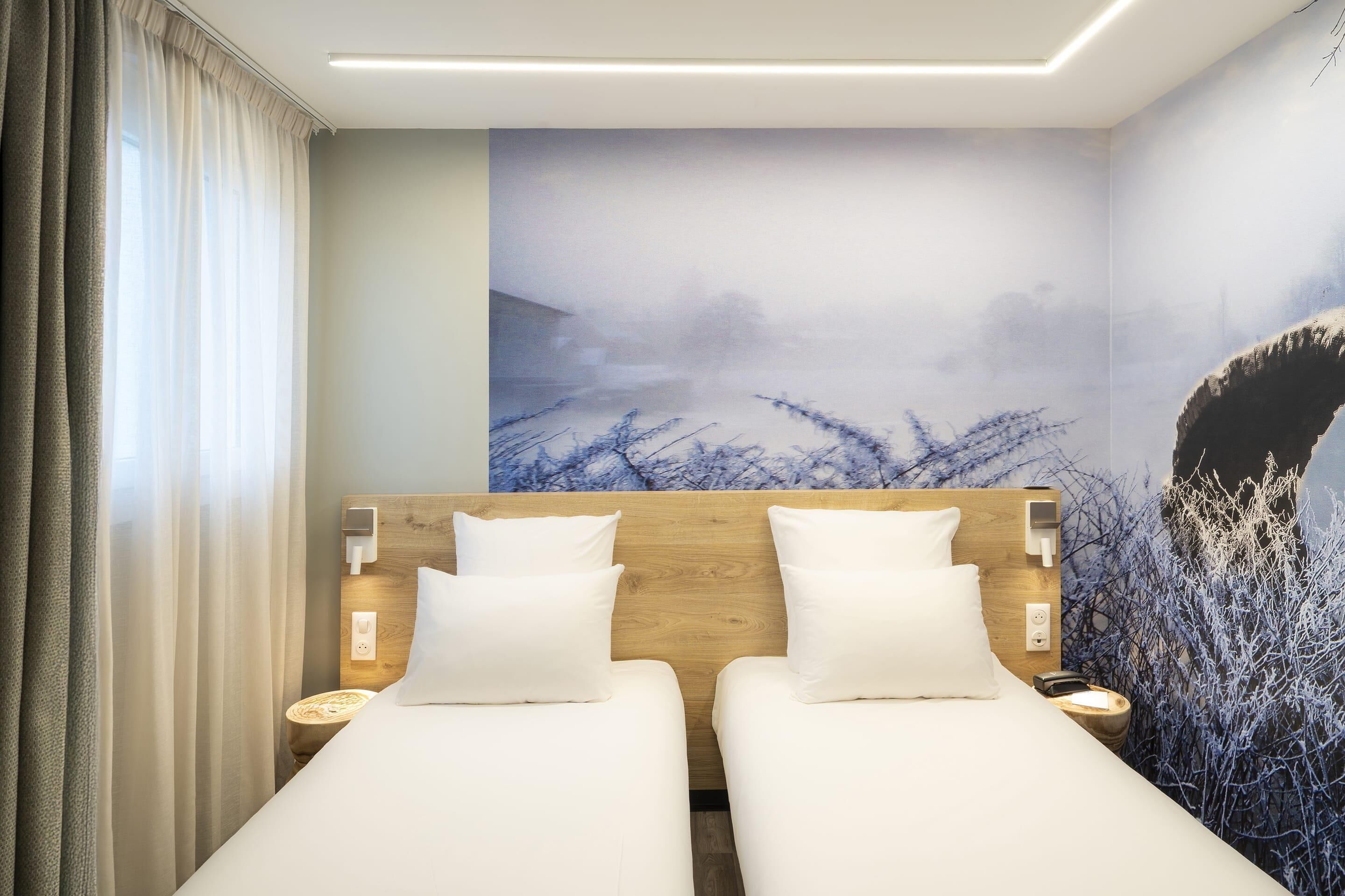 A hotel room with two beds, a wooden headboard, white bedding, a wall mural, and a bedside table with a lamp.
