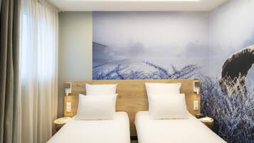 A hotel room with two beds, a wooden headboard, white bedding, a wall mural, and a bedside table with a lamp.