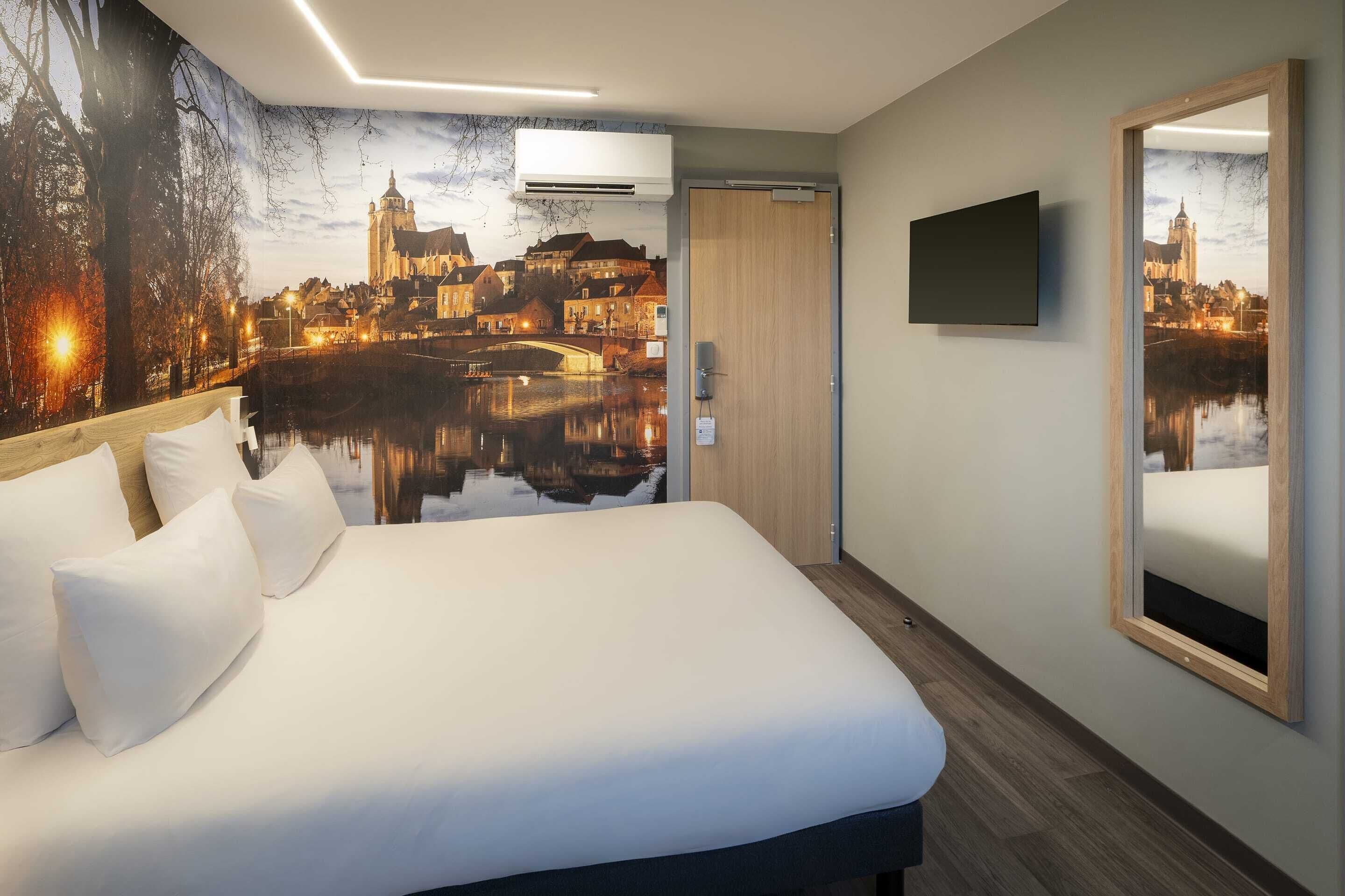 A hotel room with a large bed, a flat-screen TV, a mirror, and a cityscape mural on the wall.