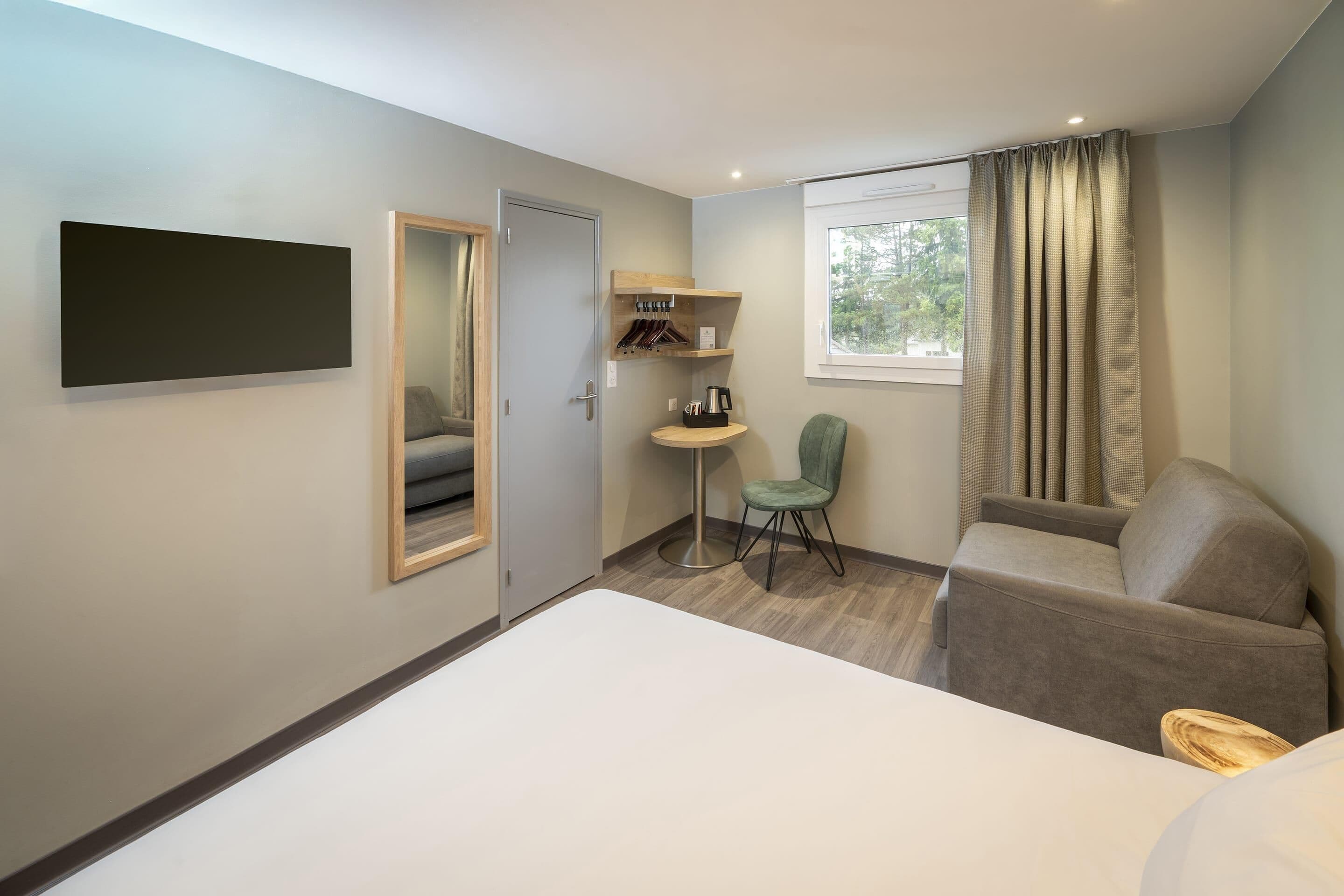 A hotel room with a bed, a seating area with a sofa and armchair, a small table with a chair, a coat rack, and a window with curtains.