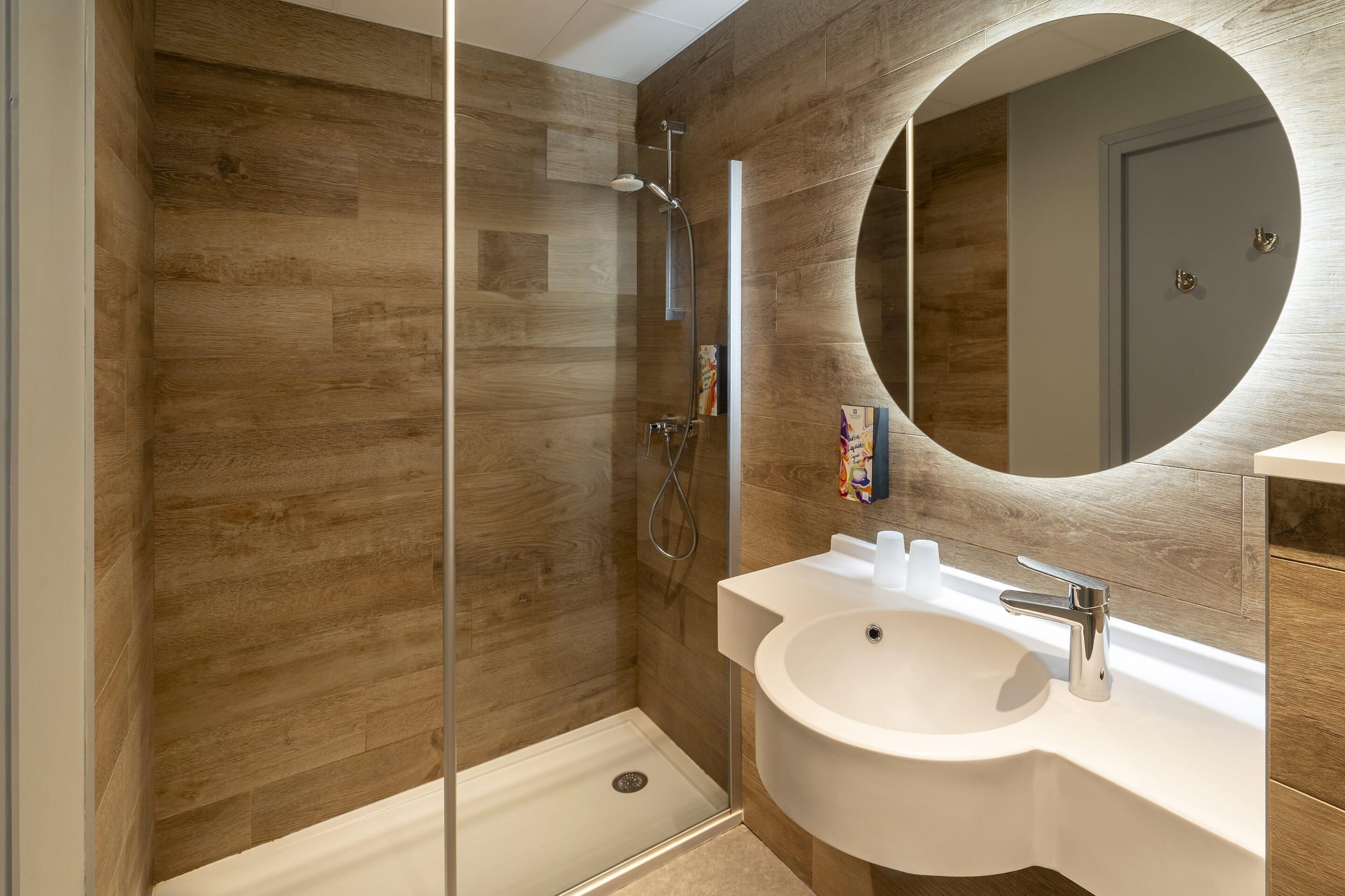 Un baño moderno con pared de madera, ducha con cerramiento de vidrio, lavamanos blanco y espejo redondo.
