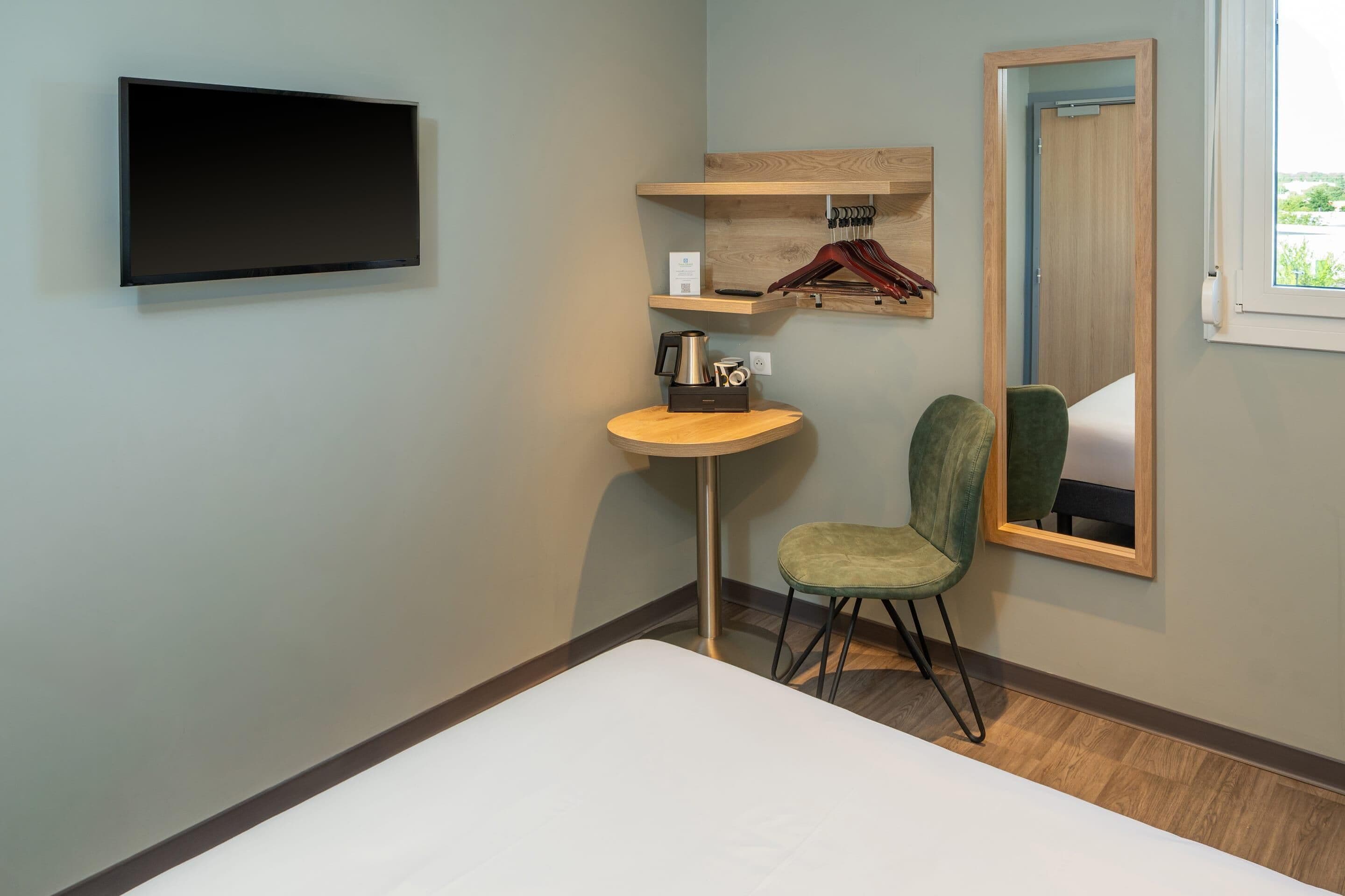Habitación de hotel con una cama, un televisor montado en la pared, una mesita redonda con una lámpara, una silla y un espejo.