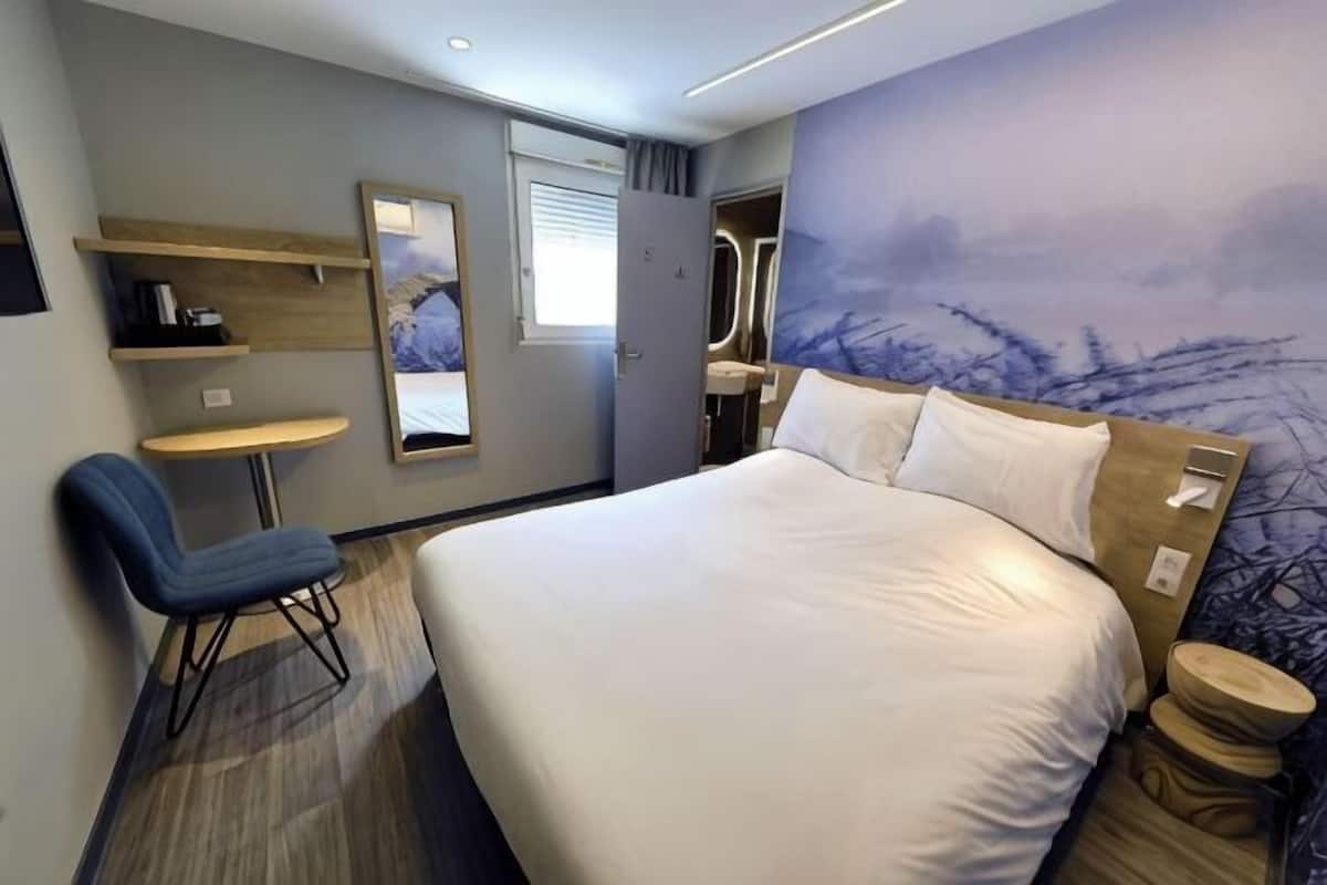 A hotel room with a bed, a desk, a chair, a small table, a mirror, and a window.