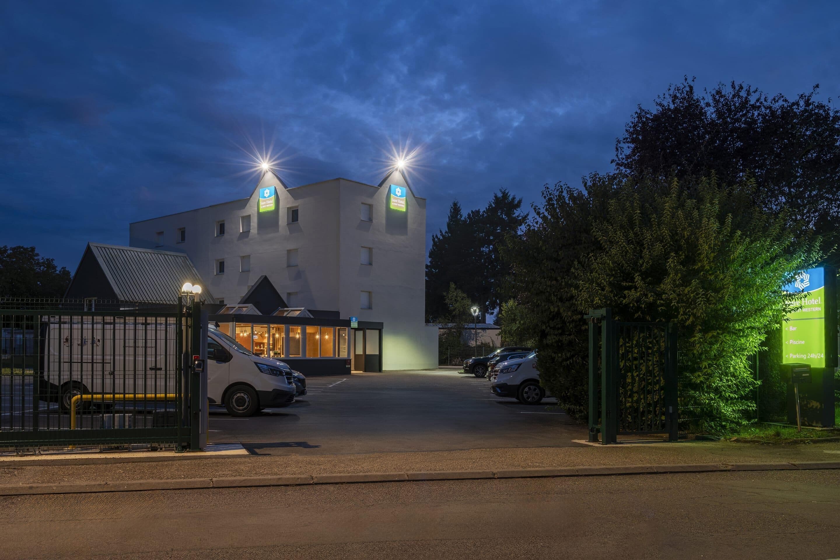 L’entrée d’un hôtel, avec une allée bordée de portails, un espace de stationnement et un panneau indiquant que le stationnement est possible 24 heures sur 24.