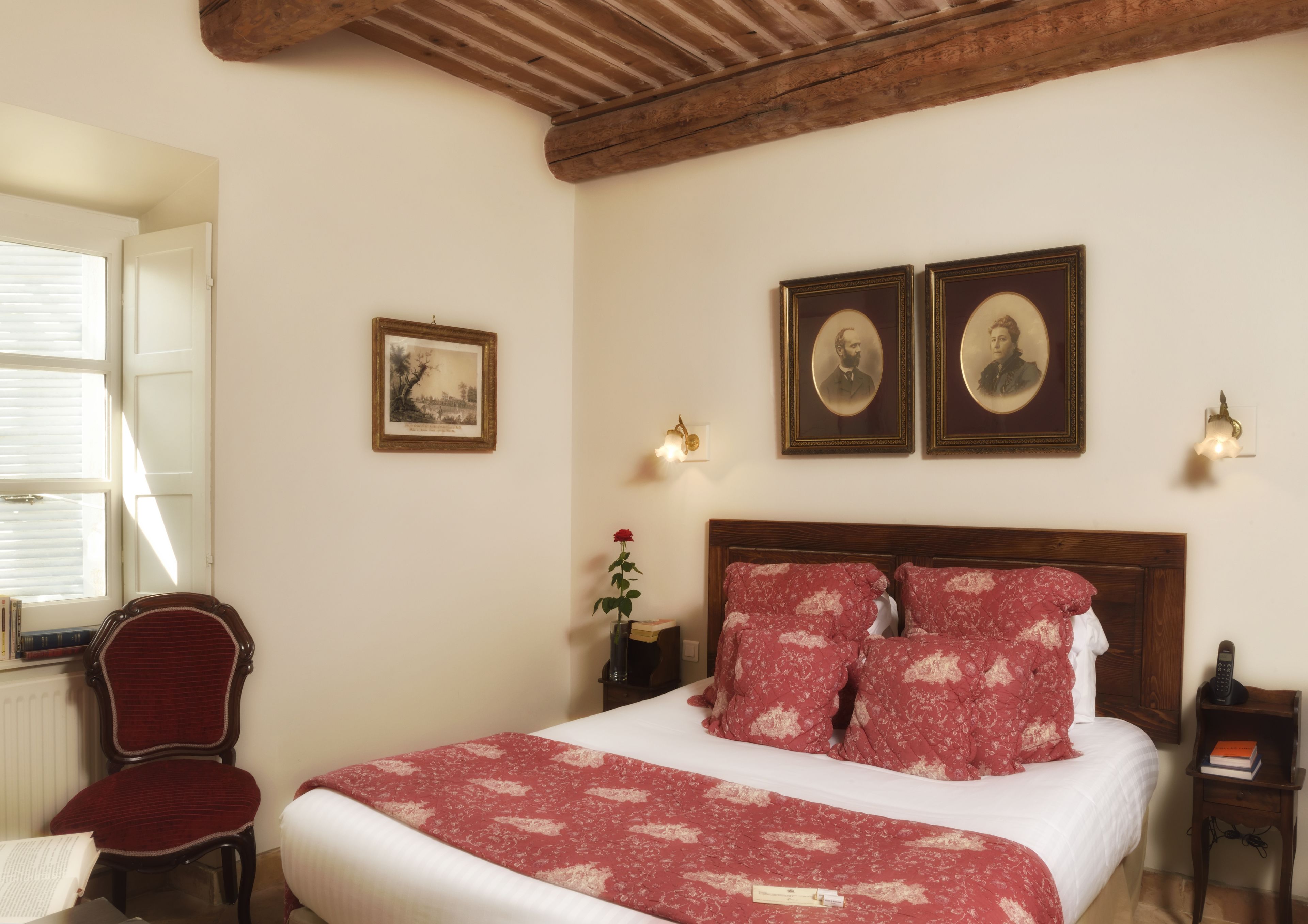 A bedroom with a wooden headboard, a bed with red floral bedding, two framed portraits on the wall, a red chair, a bedside table with a book and a vase with a single red rose, and a window with blinds.
