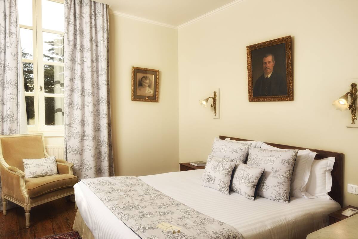 Un dormitorio con una cama grande, una silla, un ventanal con cortinas y dos cuadros en la pared.