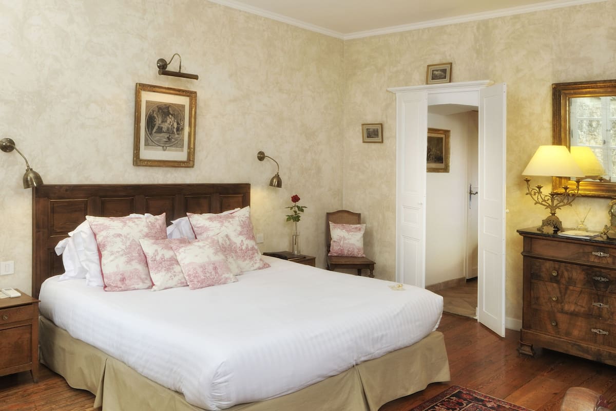Un dormitorio ordenado con una cama grande, una mesita de noche de madera, un aparador y una puerta que da a otra habitación.