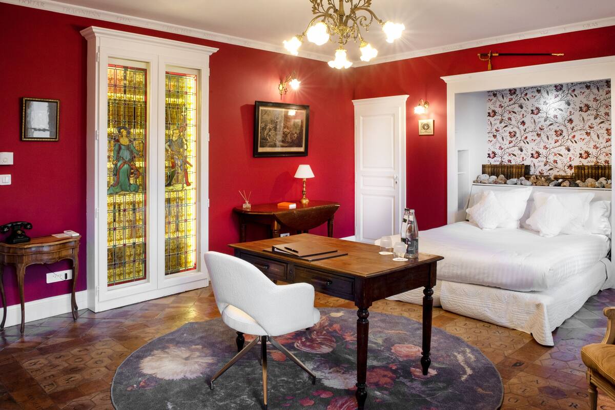Una habitación con cama, escritorio, silla, mesa, candelabro y una ventana con vitrales.