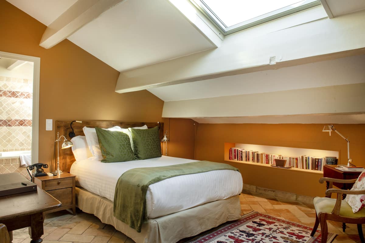 Un dormitorio con tragaluces, una cama con almohadas verdes, una mesita de noche con lámpara, una silla y una estantería llena de libros.
