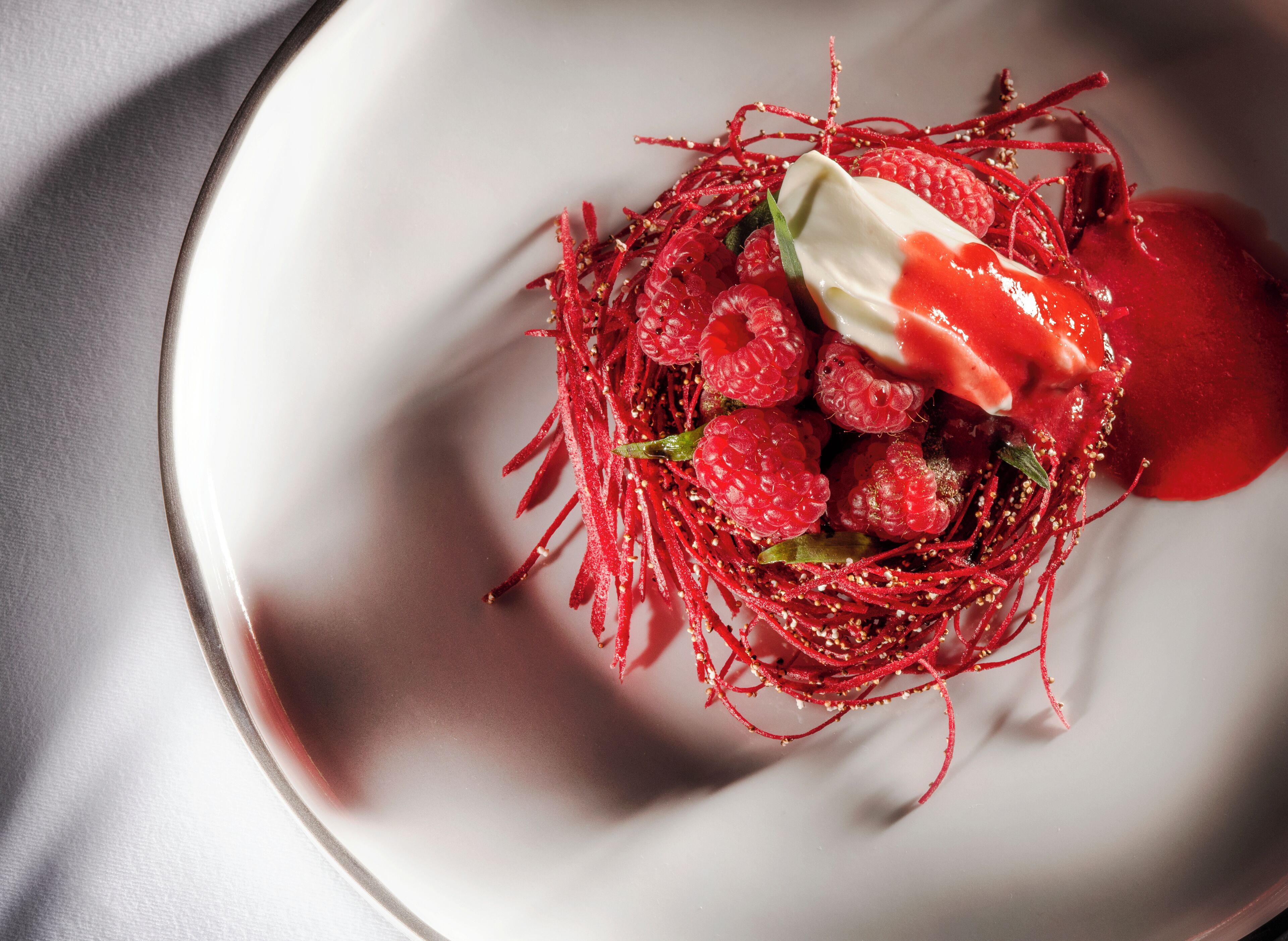 A white plate with a dessert consisting of raspberries, red threads, and a dollop of cream.