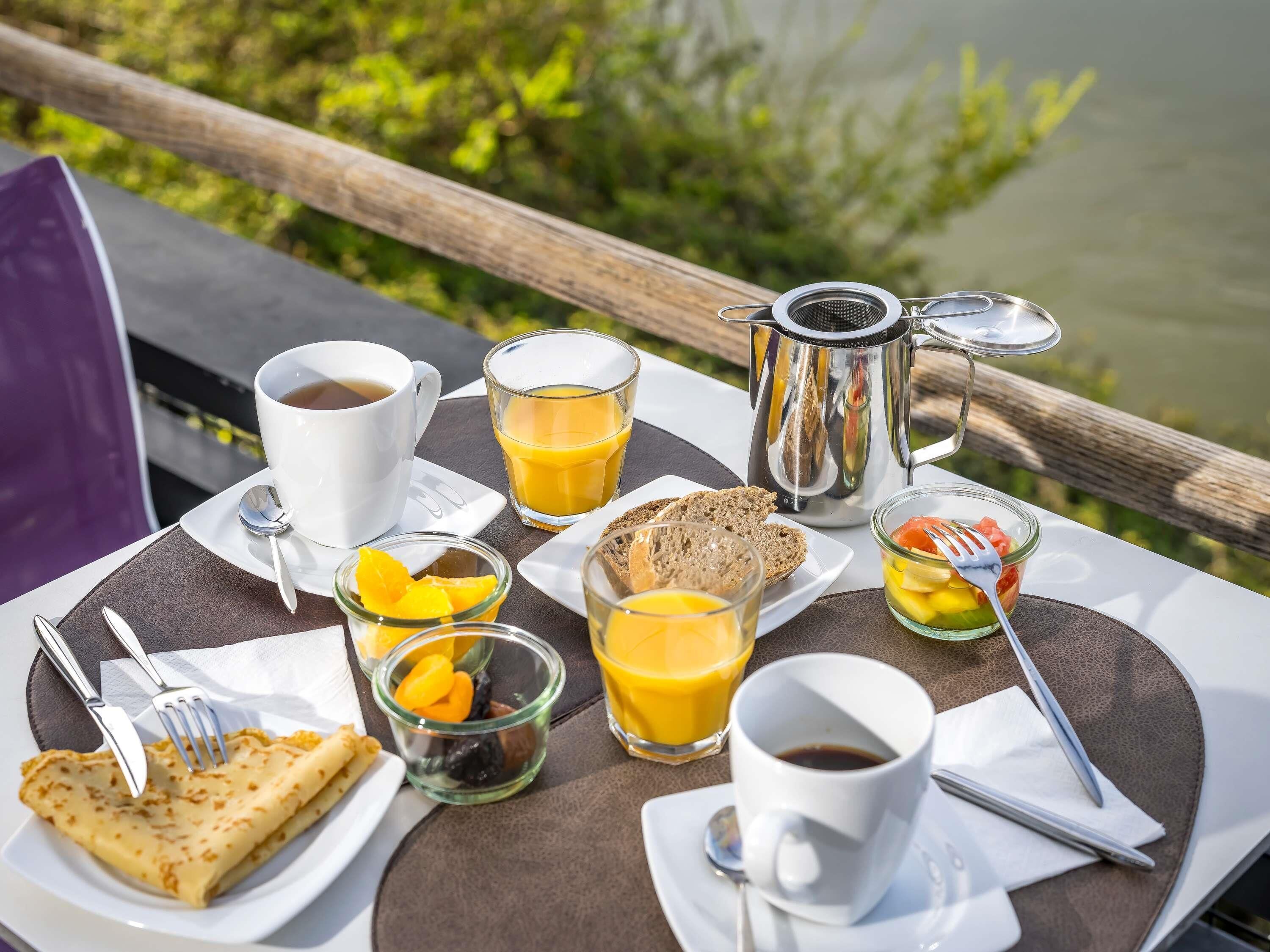 Un petit-déjeuner est préparé sur une table près d’un lac, avec du café, du jus d’orange et des toasts.