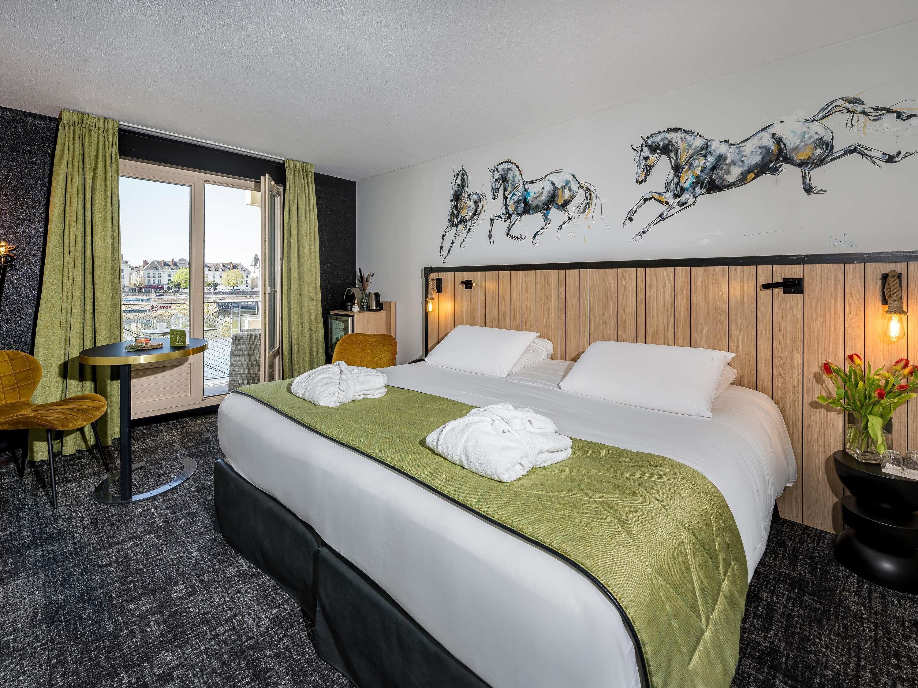 Une chambre d’hôtel comprenant un lit, un bureau, une chaise, une fenêtre avec des rideaux, un luminaire fixé au mur et un tableau représentant des chevaux.