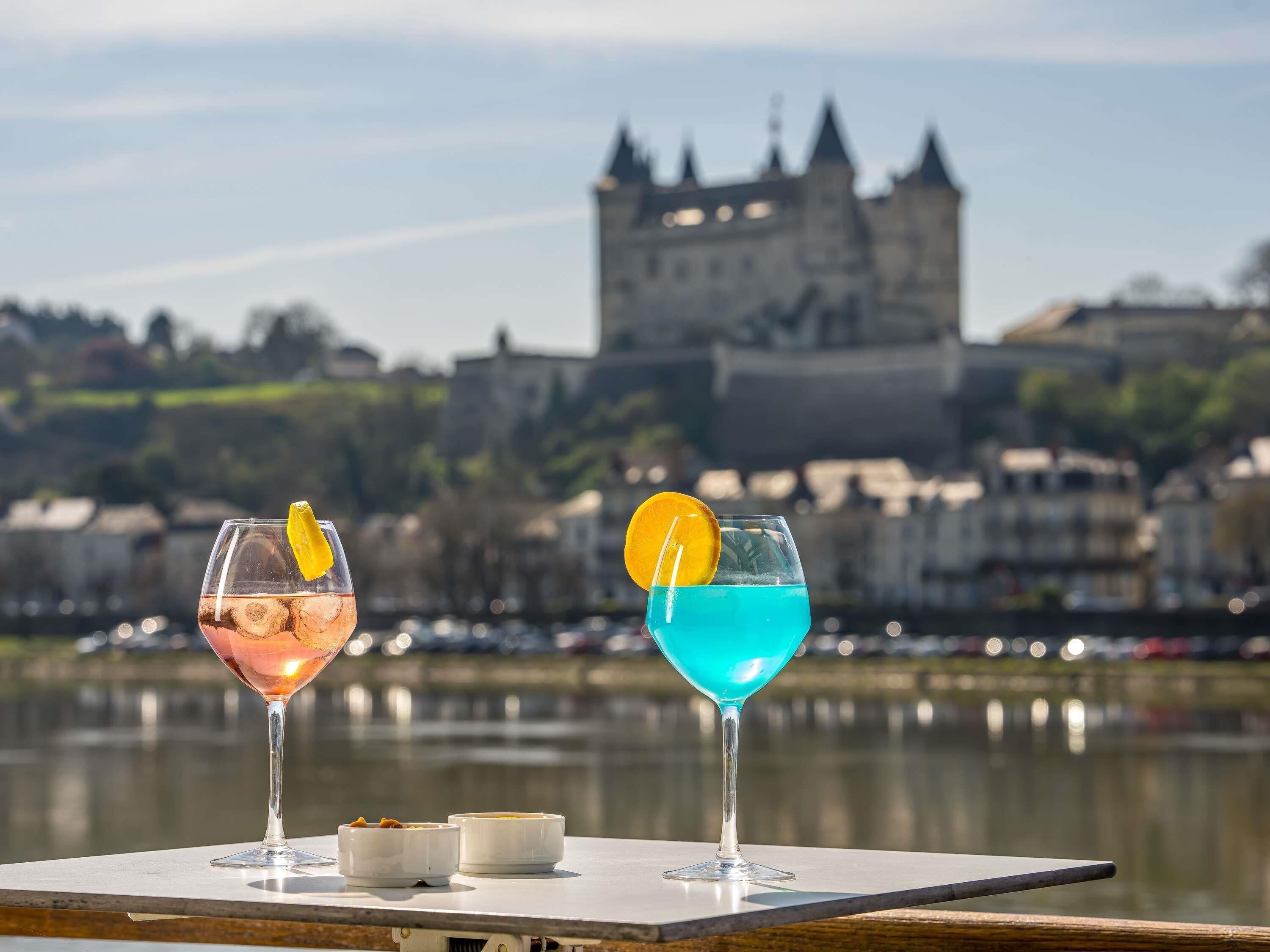 Two glasses of beverages with lemon slices on a table by a body of water with a castle in the background.