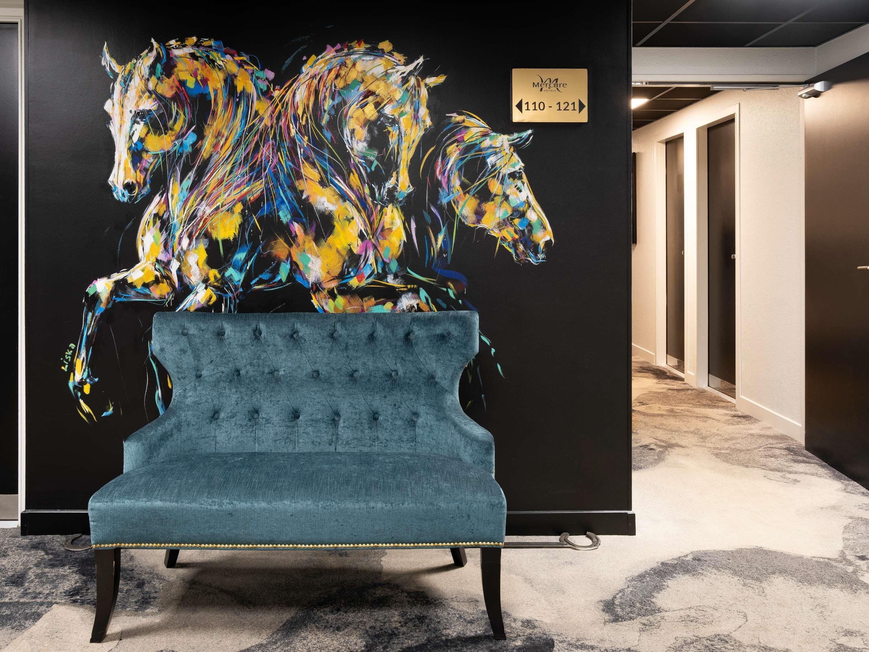 A hotel lobby with a large abstract painting of horses on a wall, a sign indicating room numbers, and a tufted blue chair in the foreground.